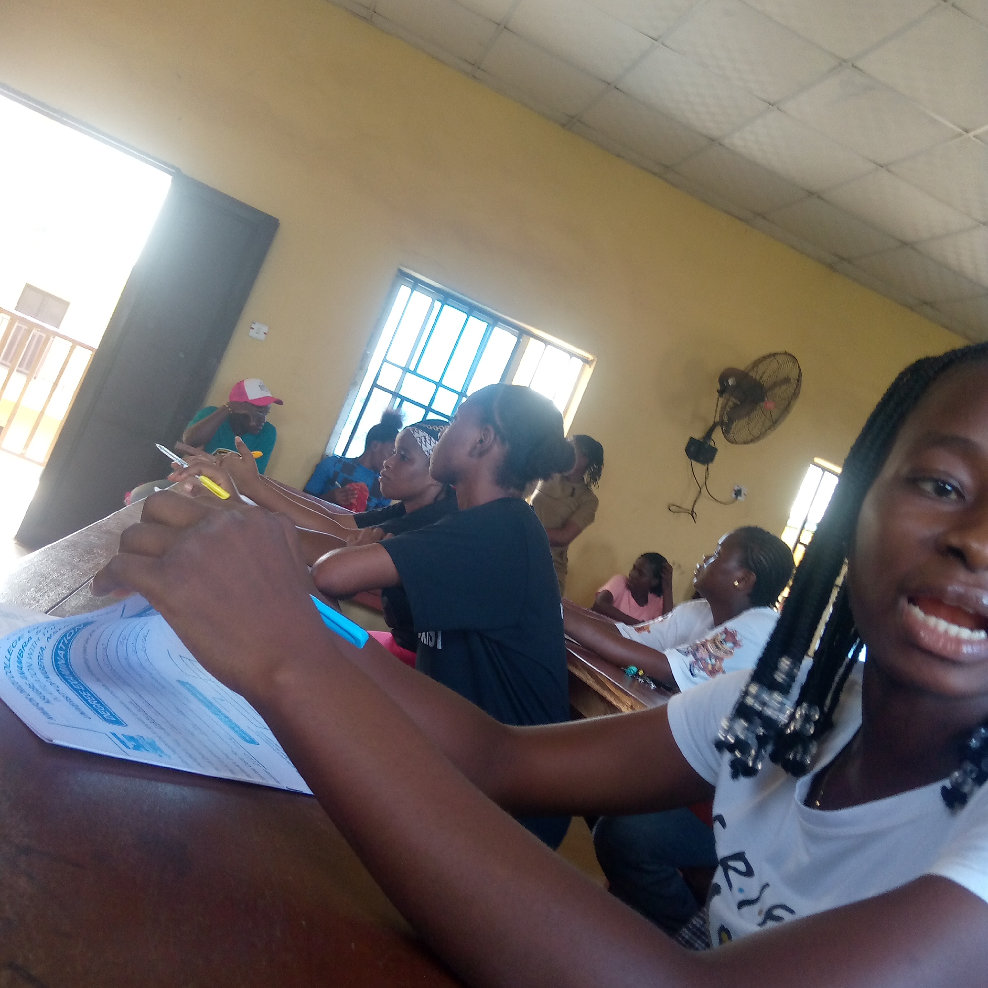 The image shows a classroom scene, likely in Ukwu-Abwa, Nigeria. Several students are seated at desks, presumably taking a test or working on an assignment. One female student is holding a blue pen while holding a paper that may be a test. In the background, other students are visible, some holding pens. The room has yellow walls, a barred window, and a ceiling fan. A doorway can be seen on the left side of the picture.