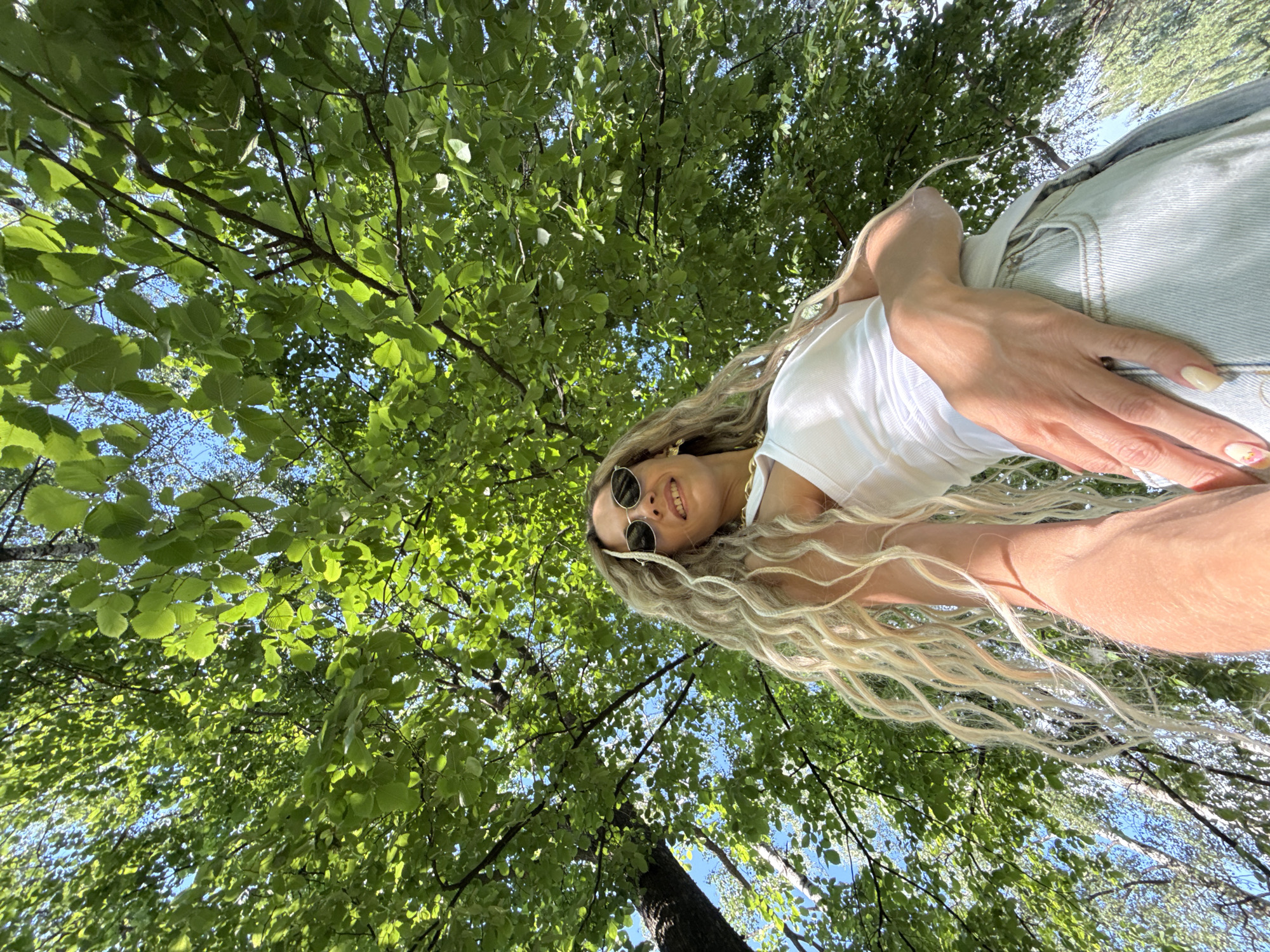 A low-angle outdoor shot shows a blonde woman wearing a white tank top, light blue denim jeans, and round sunglasses. She has long, wavy blonde hair and is wearing a chunky gold necklace. The woman is standing under a canopy of green leaves, with some of the blue sky visible through the branches. Her right arm is extended forward, with her hand resting on her leg. The image is cropped to focus on the woman and the overhead foliage. The location is identified as Rusanovo, Russia.