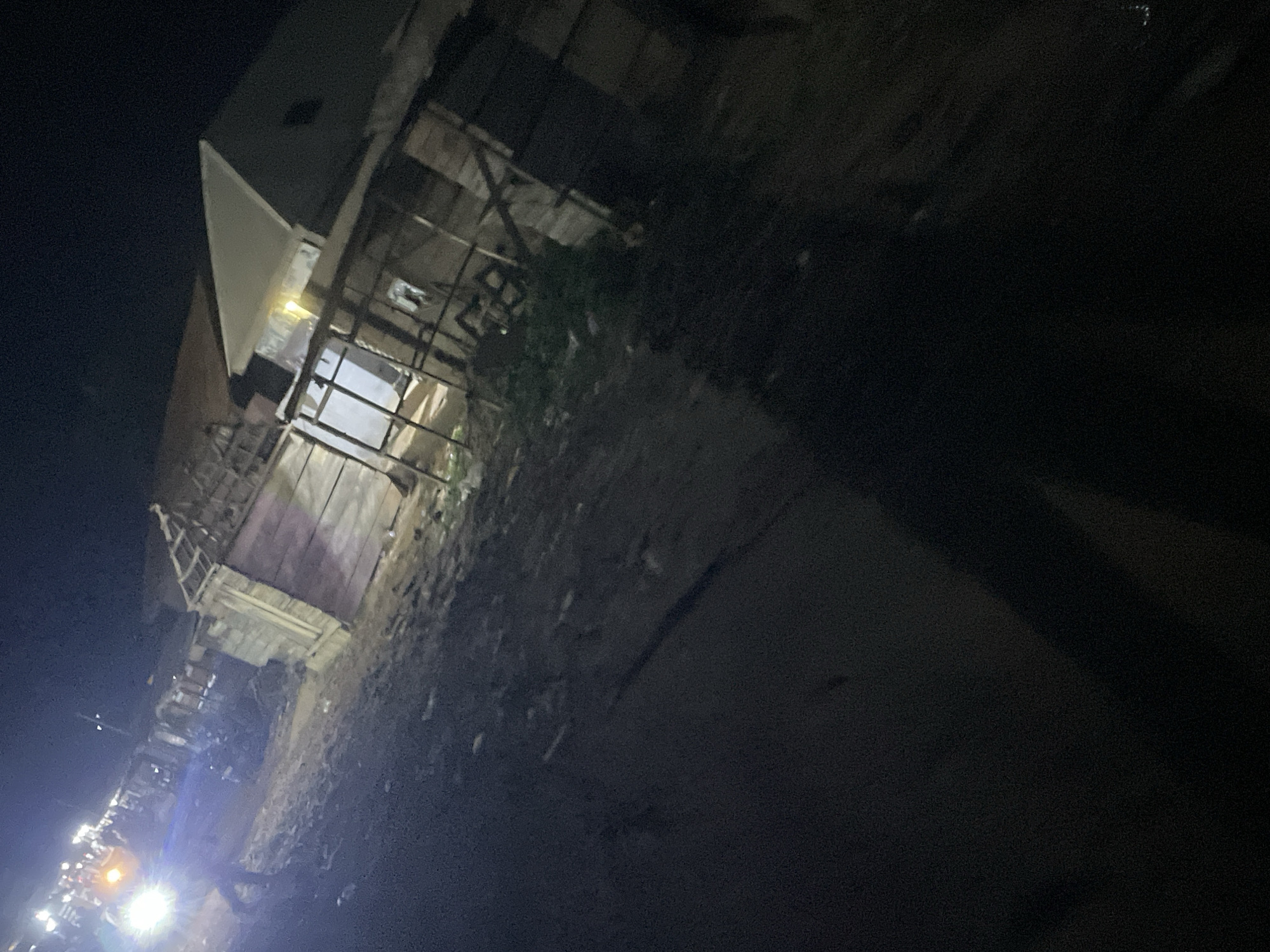This is a nighttime photograph taken in Lokoja, Nigeria, likely in an informal settlement or market area. The scene is dimly lit by artificial lights, with a bright headlight from a vehicle illuminating the left side of the image. A person is visible walking in the foreground, partially silhouetted by the vehicle's light.
The background features a row of structures that appear to be makeshift shops or dwellings, constructed from wood and corrugated metal. Some of these structures are incomplete or in disrepair, with exposed wooden beams and framing. One structure in the center has an open doorway, with light spilling out. The ground is uneven and appears to be dirt or gravel, with debris scattered around. The overall impression is one of a busy but potentially impoverished urban environment at night.