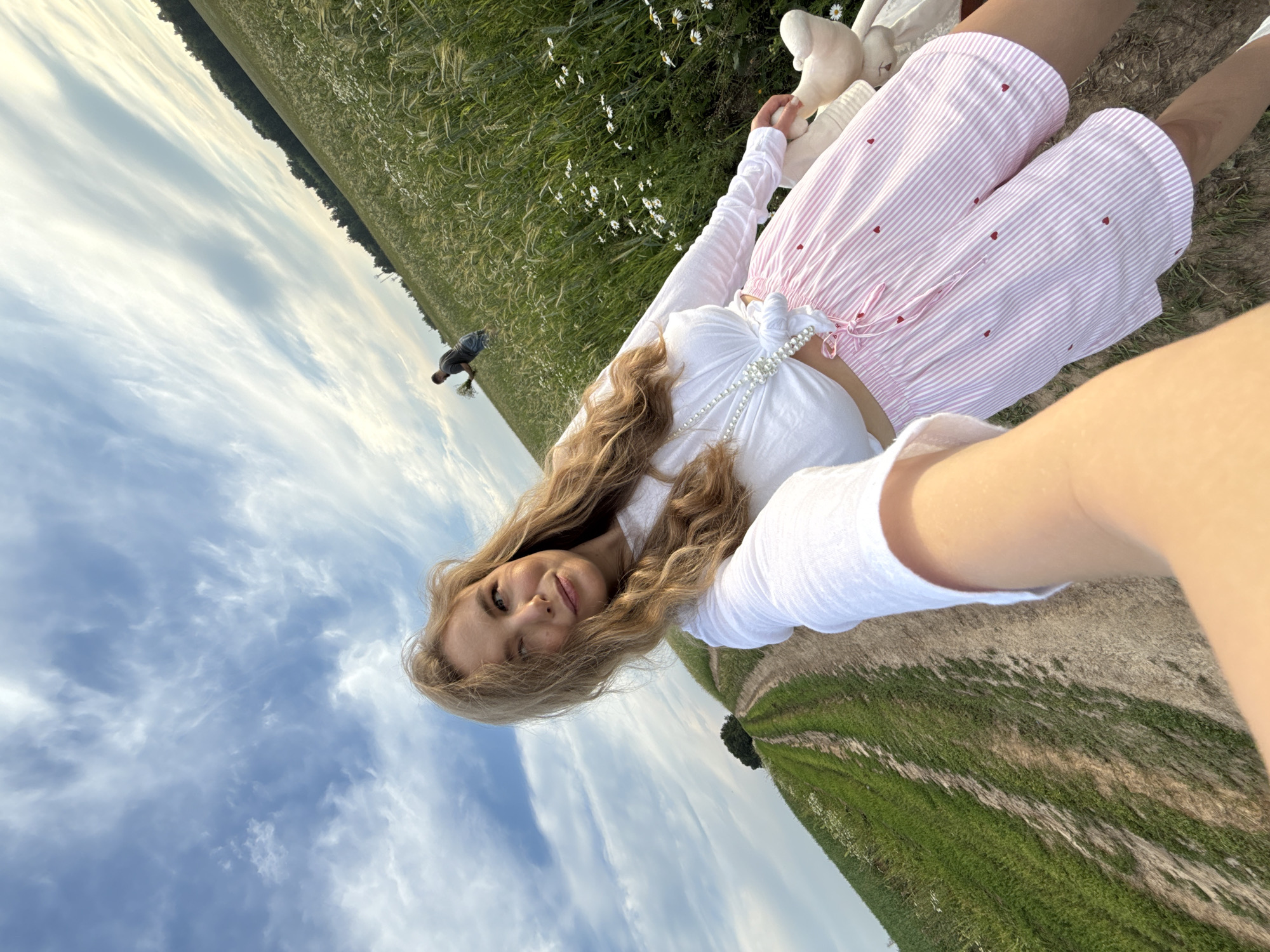 An outdoor, low-angle, selfie-style shot captures a young woman in a field. She has blonde hair, is wearing a white cropped top and pink striped shorts with red hearts, and is holding a stuffed animal. To her right is a field of green grass with white flowers and trees in the background. A dirt road with green grassy edges is to her left. In the distance, a man is walking through the field, carrying a bouquet of flowers. The sky above is blue with wispy white clouds. The location is specified as Сновицы, Russia.