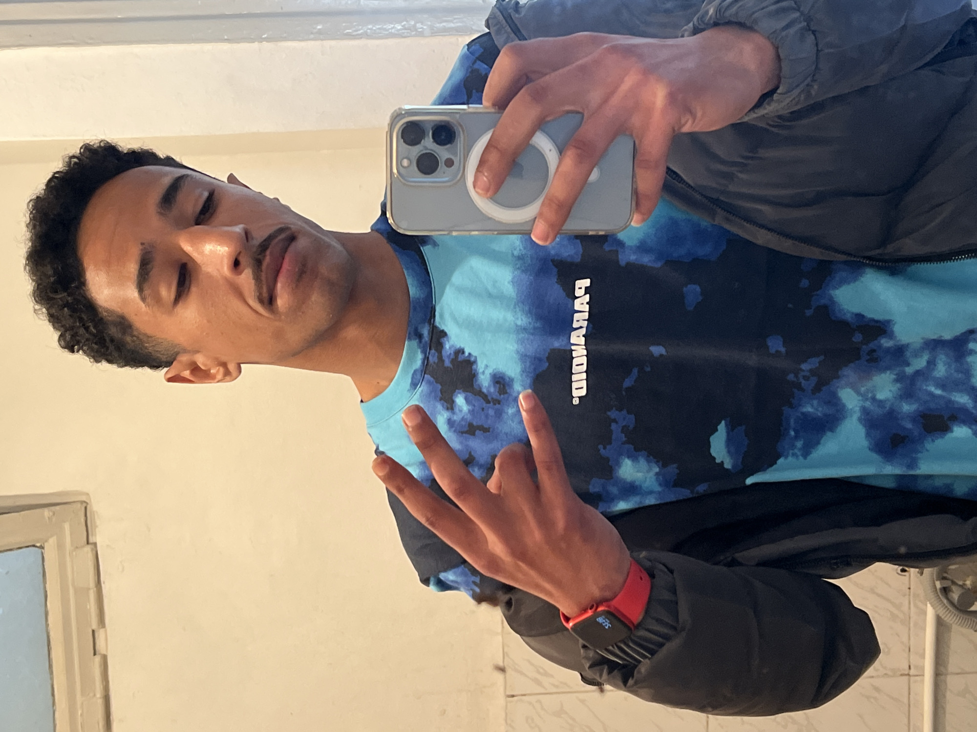 A man is taking a selfie in what appears to be a bathroom. He is wearing a blue tie-dye shirt with a black jacket over it, and a red smartwatch on his left wrist. He's holding a phone with a clear case. The background shows a plain off-white wall with a window. There is no evidence of the location being in Tanger, Morocco.