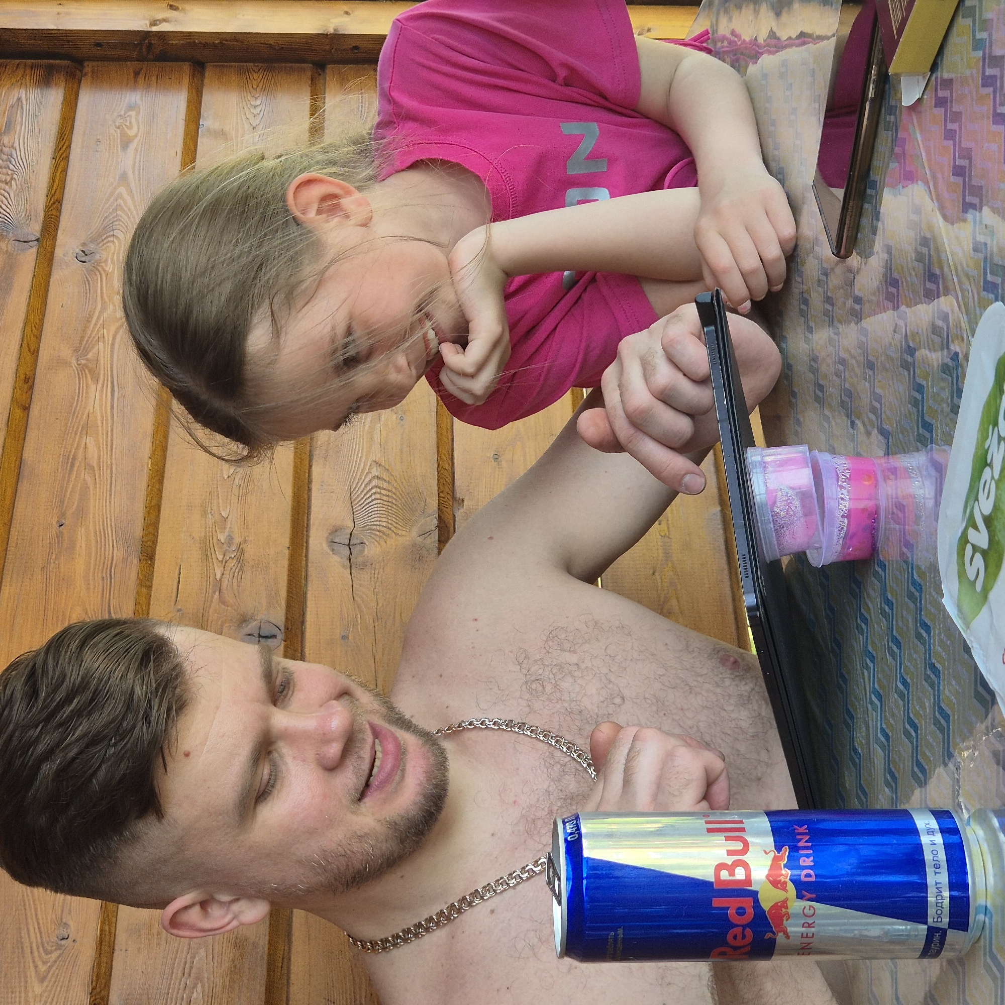In Новостройка, Russia, an adult male and a young female are seated at a table against a background of horizontal light-brown wooden planks. The male, with short dark hair and a beard, is shirtless but wears a gold-colored chain necklace. He looks down at a black tablet, pointing at its screen with his right index finger, while his left hand holds an open blue and silver Red Bull energy drink can. The can displays Russian text including "Бодрит тело и дух" (Invigorates body and spirit). To his right, a young female with light-brown hair, wearing a bright pink t-shirt with a silver "ON" visible, smiles broadly while observing the tablet screen, her chin resting on her left hand. The tablet is propped on two stacked, clear plastic containers filled with a pink, possibly glittery, substance. The table is covered with a reflective, patterned tablecloth featuring geometric designs in blue, green, yellow, and white. A dark-colored smartphone and a small yellow-brown cardboard box are also on the table, along with a partially visible white item with green text at the bottom left.
