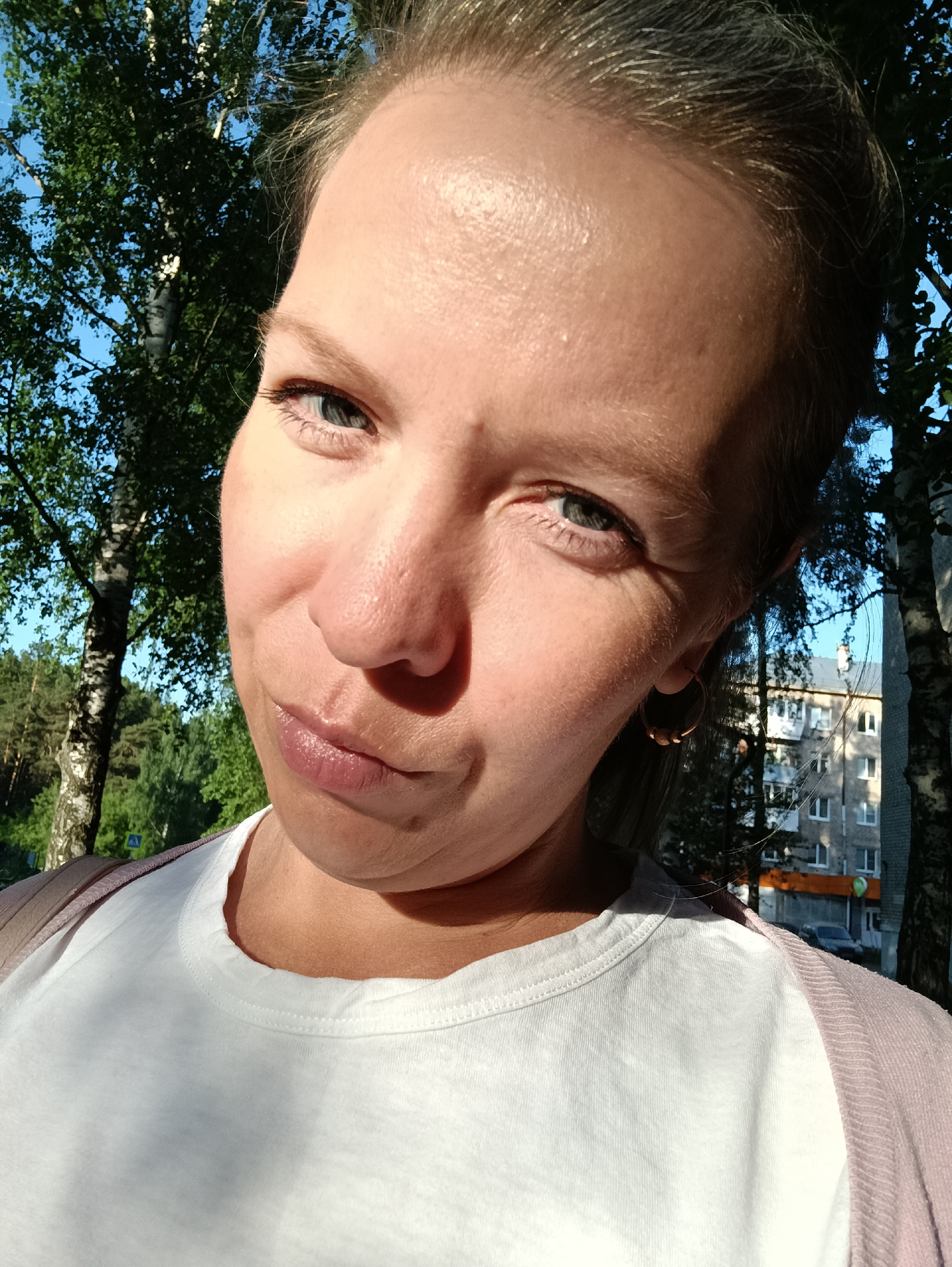 A close-up selfie of a fair-skinned woman with light-colored hair pulled back, posing outdoors on a sunny day. She is wearing a white t-shirt and a light pink sweater. Green trees are visible in the background, along with a multi-story building in the distance with visible windows. The building has a sign in Russian, indicating it's likely in Russia. Based on a prior question and the presence of birch trees, the location could be Chaikovsky, Russia. The woman has light eyes and a slight smirk.