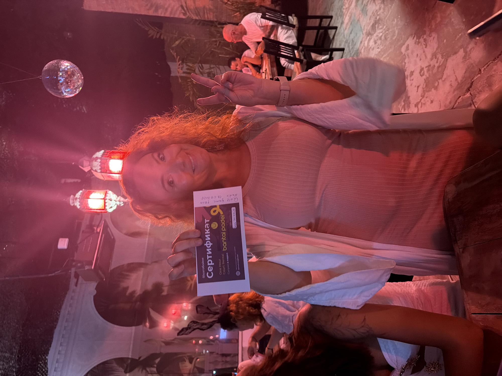 This media file depicts a woman holding a certificate and smiling at the camera, with her hand raised in a peace sign. The certificate, which appears to be a "Quiz game FROM Nadir" from "bantai padel," is in Russian and mentions "One Hour Court Rental." The scene is dimly lit with pink and red hues from overhead lighting, including a disco ball and decorative lanterns. In the background, other people are visible sitting at tables. The overall atmosphere suggests a social gathering or event, possibly at a venue with a dance floor or entertainment area, given the disco ball. The woman's expression is cheerful and engaging. The presence of the certificate and the reference to a "quiz game" and "court rental" indicate some form of organized activity or entertainment. The lighting and decor suggest an evening or night time setting. The location context provided places this scene in Ban Rak, Thailand.