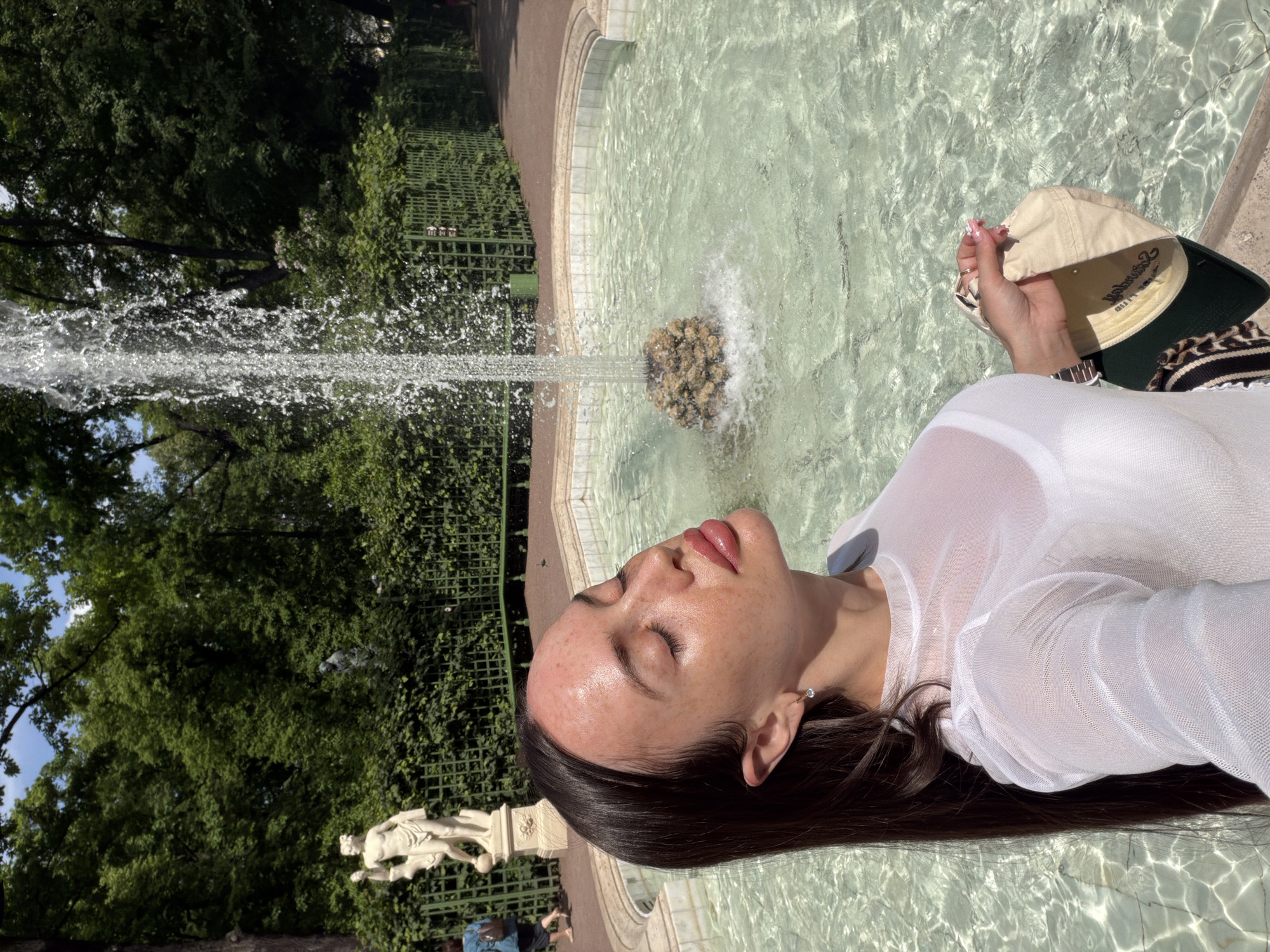 A young woman with dark hair and her eyes closed tilts her head back to feel the sun, standing next to a fountain in a park. She is wearing a sheer white top over a white bra. In her right hand, she holds a tan baseball cap with a green underside. The fountain has clear, shallow water and a decorative rock in the center. Behind the fountain, there is a green metal fence and manicured bushes. To the left of the fountain, a white statue of a male figure stands on a pedestal. The background is filled with lush green trees. The location is the Summer Garden in Saint Petersburg, Russia.
