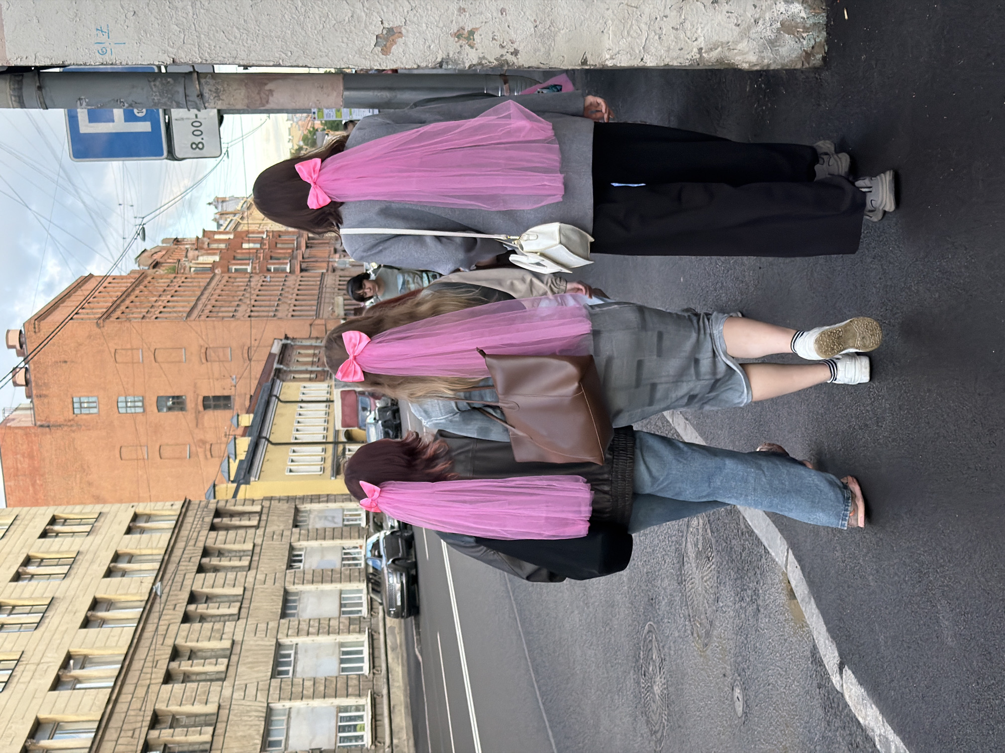 Three individuals, seen from behind, walk along a sidewalk in Saint Petersburg, Russia. Each individual has a large pink bow attached to their hair, with a sheer pink veil trailing down their back. The person on the left wears a dark jacket, blue jeans, and pink sandals. They carry a black bag. The person in the middle wears a denim jacket, a grey skirt, and white sneakers with black and white striped socks. They carry a brown leather tote bag. The person on the right wears a grey blazer, black pants, and grey sneakers. They carry a white handbag. They are walking away from the viewer on a paved sidewalk, which is wet in places. Across the street are buildings with classical architecture, typical of Saint Petersburg, and some newer brick buildings. A blue parking sign with the letter "F" is visible on a pole on the right side of the image. The sky is overcast.