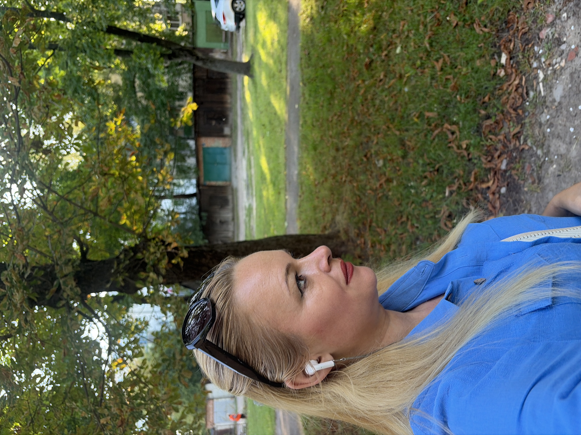 An outdoor, profile shot shows a blonde Caucasian woman in her late 30s or early 40s, wearing a blue collared shirt and black sunglasses perched on her head. She has on white earbuds. She is looking to her right. Her hair is pulled back. The woman is in front of a grassy area with some fallen leaves. Behind her are trees, some with green leaves and some with yellowing leaves, and in the distance are some garages and a white car.