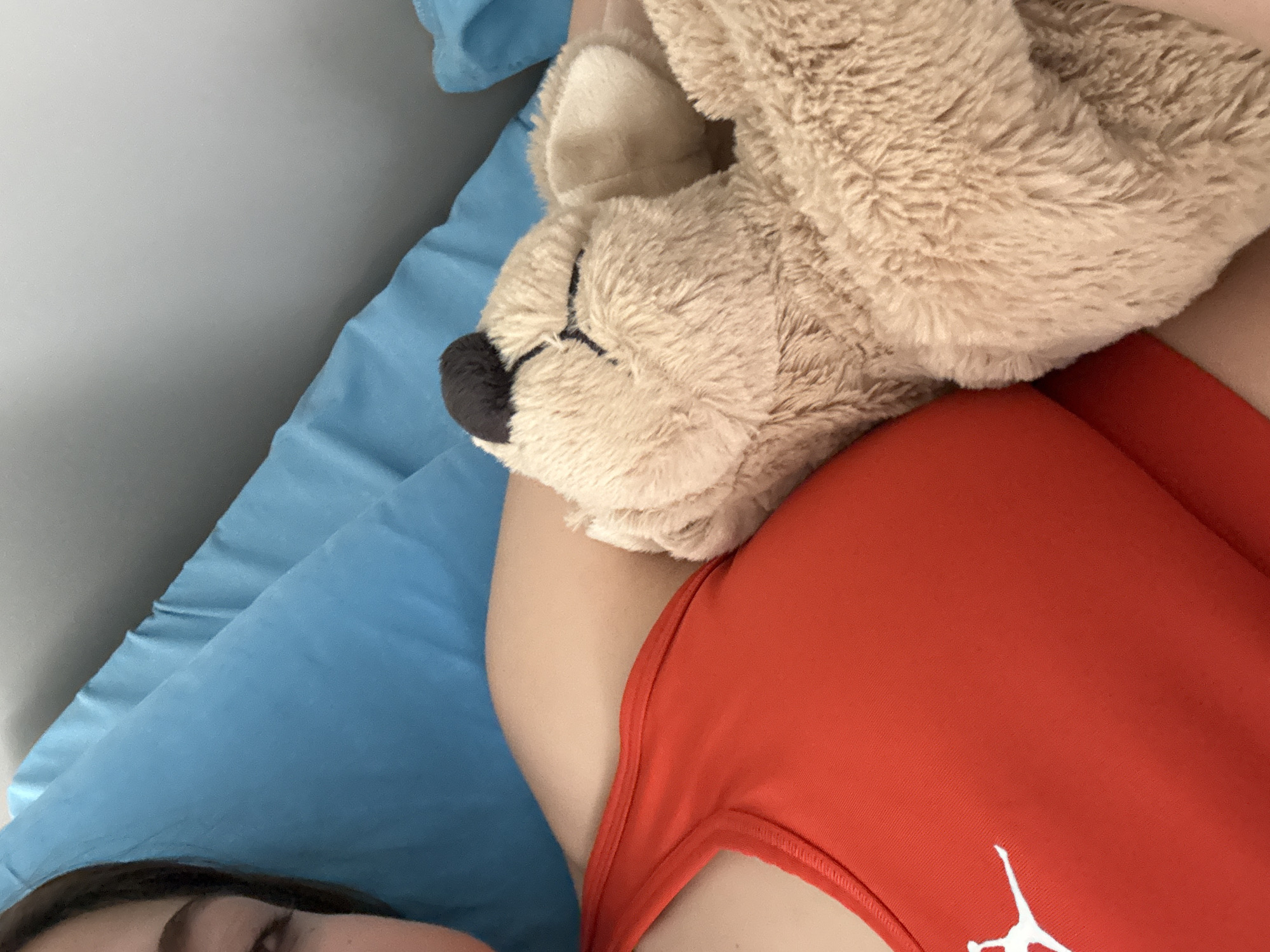 A person wearing an orange tank top lies in bed with a stuffed animal. The person is on a blue pillow, and only part of the face is visible. The stuffed animal is beige with a black nose. The location is not discernible from the provided image.