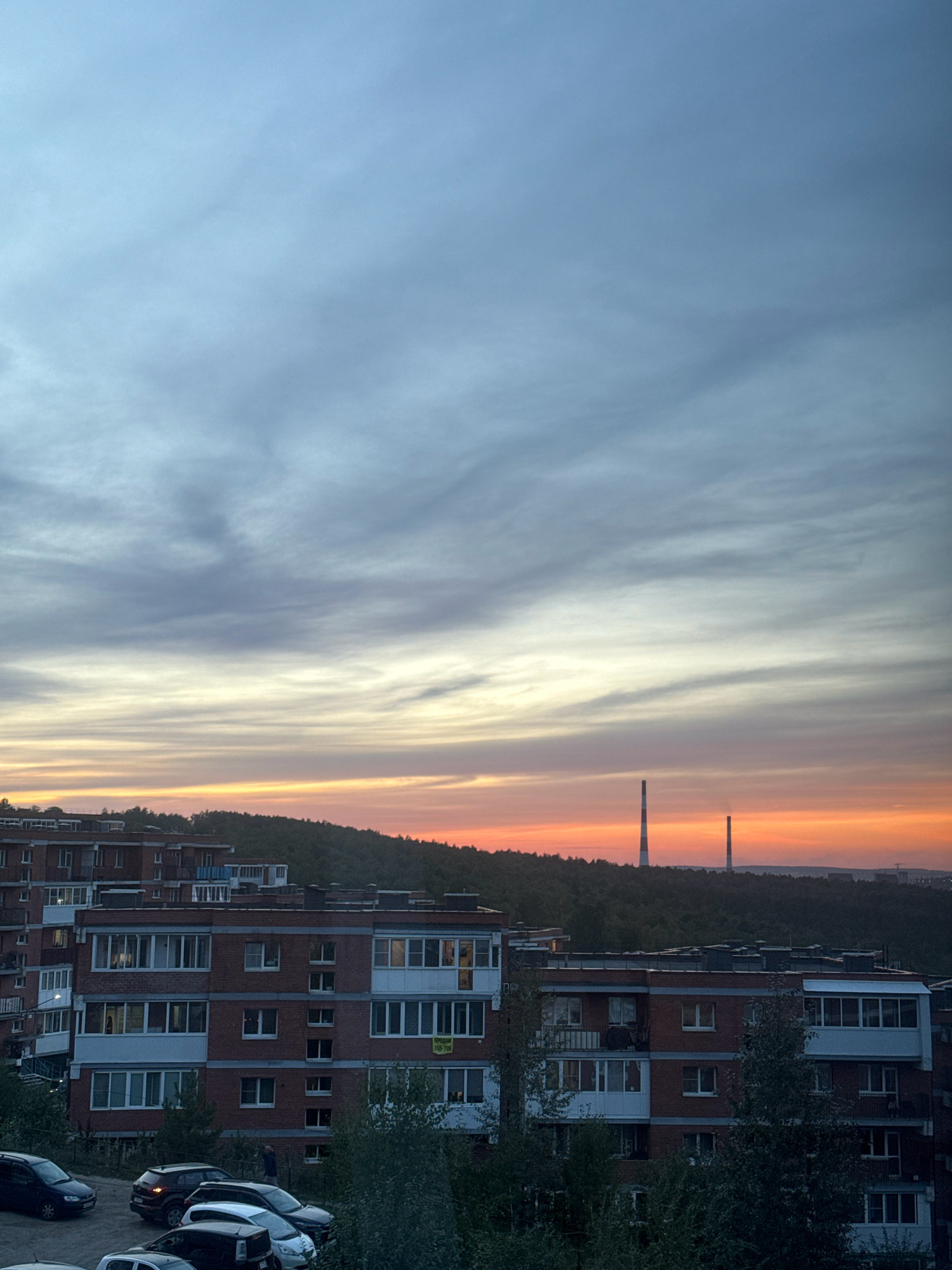 The image depicts a residential area in Markova, Russia, during a sunset. In the foreground, several apartment buildings with red brick facades and numerous windows are visible. Cars are parked in a lot in front of the buildings, and trees and vegetation surround the structures. In the background, a dense forest covers rolling hills, and two tall, industrial smokestacks stand prominently against the colorful sky. The sky itself displays a gradient of colors, transitioning from pale blue at the top to vibrant orange and yellow near the horizon, with wispy clouds scattered throughout. The overall scene suggests a late evening in a suburban or semi-urban environment.