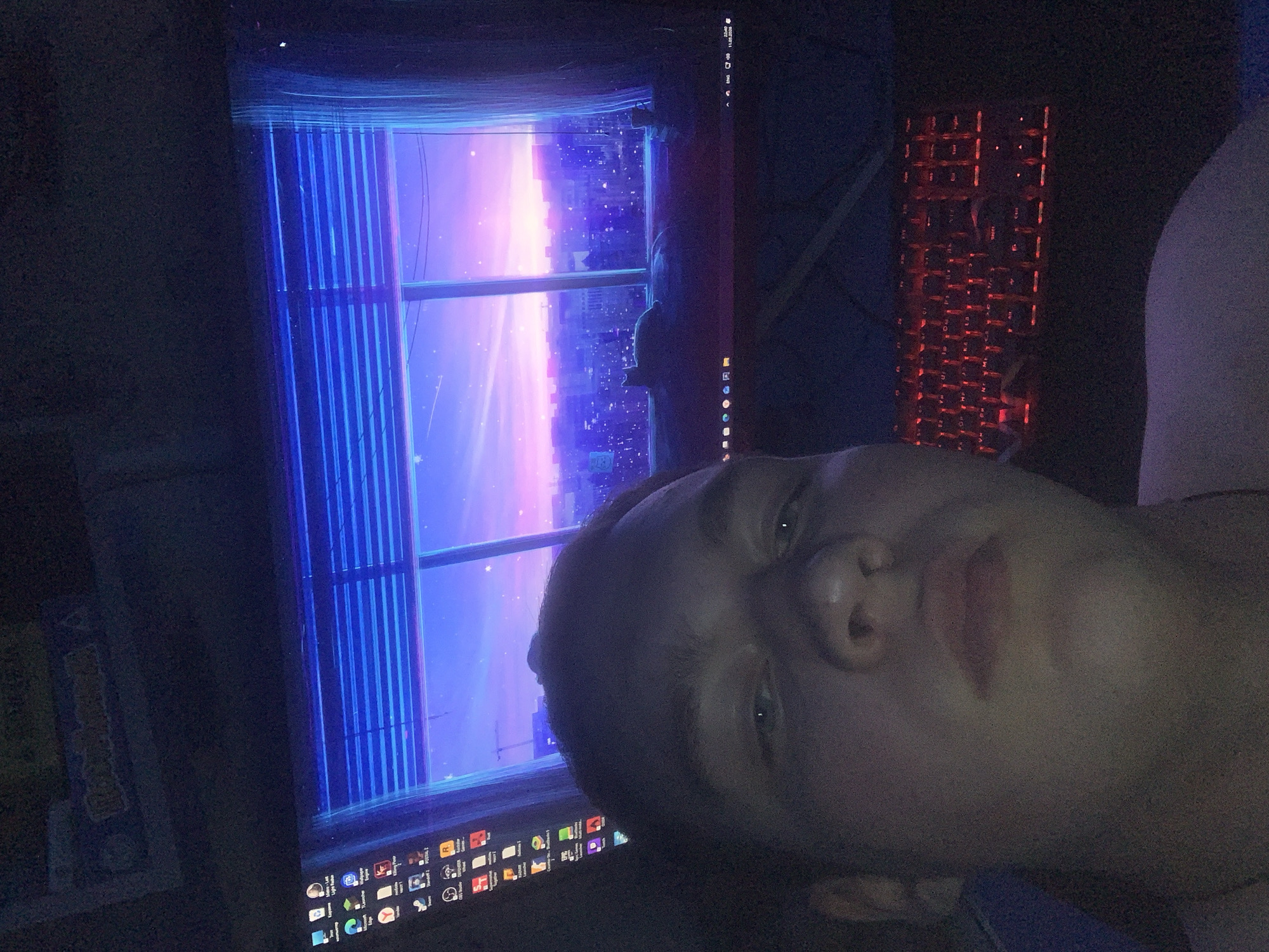 A person's face is prominent in the foreground, oriented at an angle towards the camera. Behind the person is a computer monitor displaying a cityscape view with purple and blue hues resembling a sunset or dawn scene. The screen also shows a blue, barred window frame. To the right of the monitor is a partially visible red backlit keyboard. The scene appears to be indoors and dimly lit. The city of Severodvinsk, Russia, cannot be confirmed as the location based on the visible content of this image.