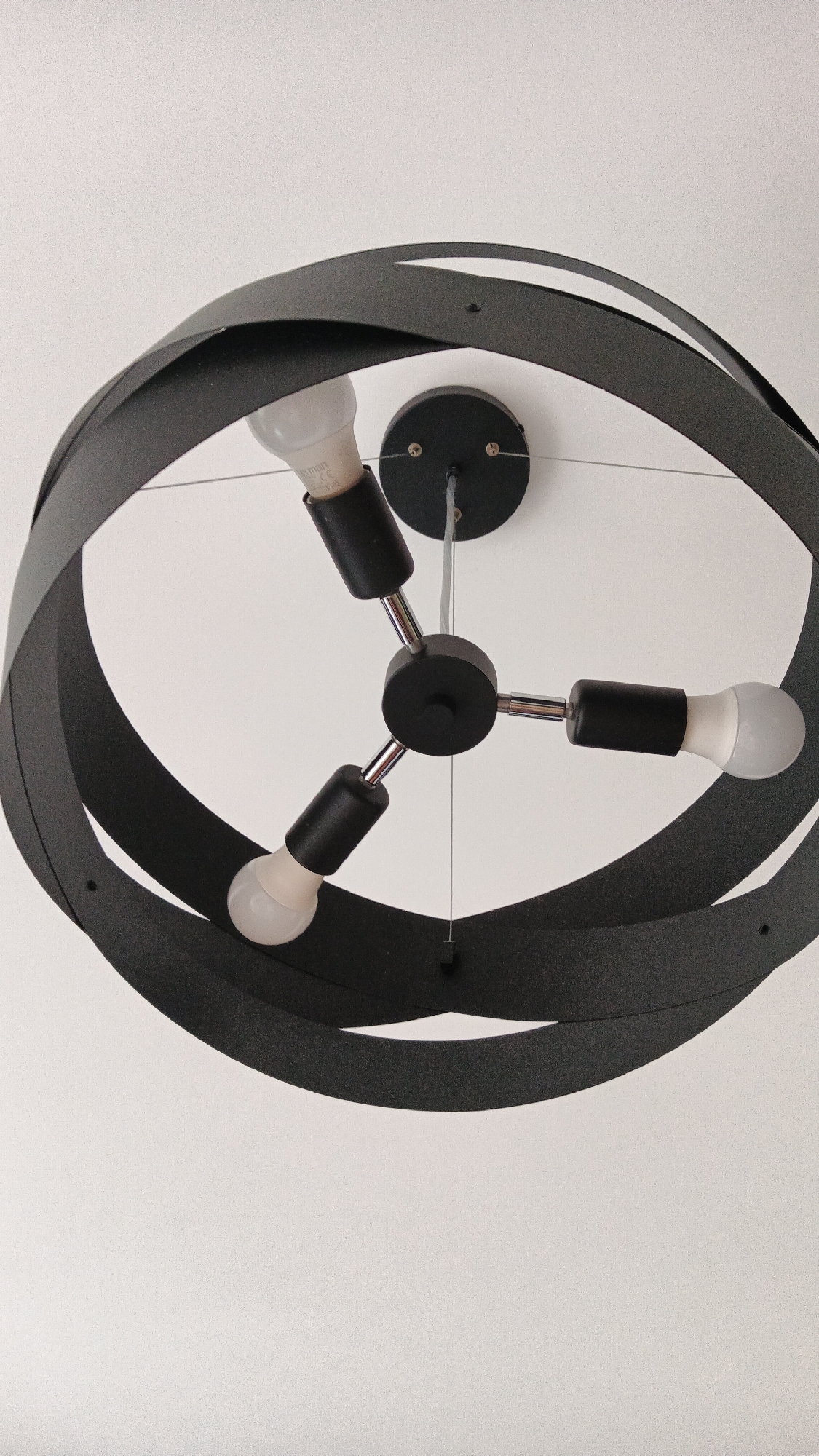 A contemporary, black, circular chandelier hangs from a white ceiling. The chandelier has a twisted, ribbon-like design that forms an outer ring. Inside the ring, a central black fixture with three metallic arms extends outwards, each holding a black socket with a white LED bulb. The bulbs are frosted and visible, with some text printed on them. The chandelier is suspended by thin wires from a black circular ceiling mount. The scene is brightly lit, with a slight graininess to the image. There are no people or specific environmental features visible besides the ceiling and the chandelier. No specific location in Zielona Góra, Poland, can be identified from the image.