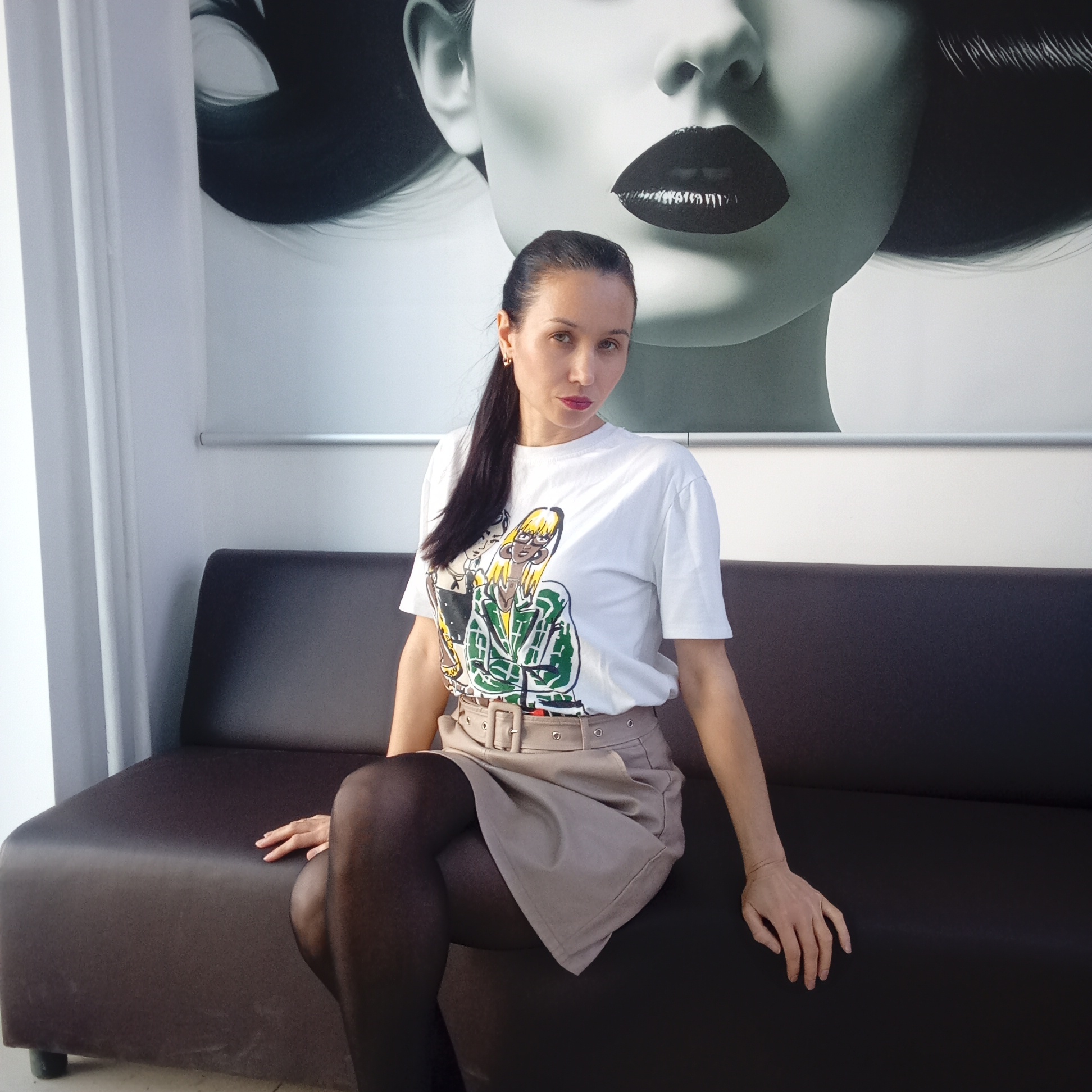 A woman is sitting on a dark brown couch in an indoor setting. She has long, dark hair pulled back into a ponytail, and she is wearing a white t-shirt with a cartoon design, a tan skirt with a belt, and black tights. Her legs are crossed. Behind her is a large black and white image of a woman’s face, featuring pronounced dark lips. To the left of the scene, there is a white curtain, and above the couch, there is a silver rail on the wall. The environment appears to be a room in Izhevsk, Russia, as indicated in the prompt. The woman is posing for the photograph.