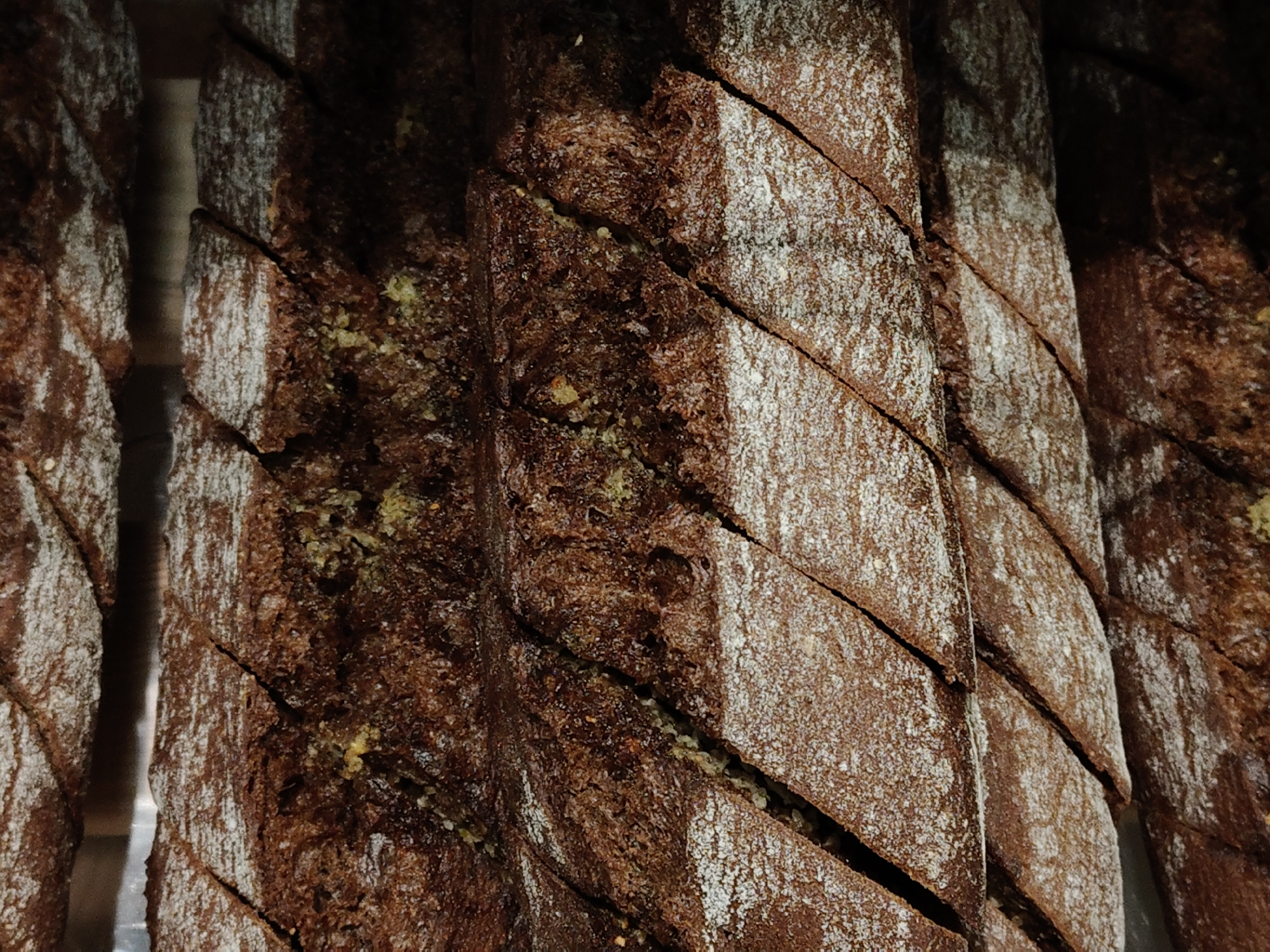 A close-up shows several sliced loaves of dark bread, possibly rye or pumpernickel, arranged vertically. The bread is crusty, with a dusting of what appears to be flour on the cut surfaces. The slices are separated by narrow gaps. Patches of what might be green mold are visible on one of the loaves. The scene appears to be inside a bakery or food market. Location: Unknown, Unknown.