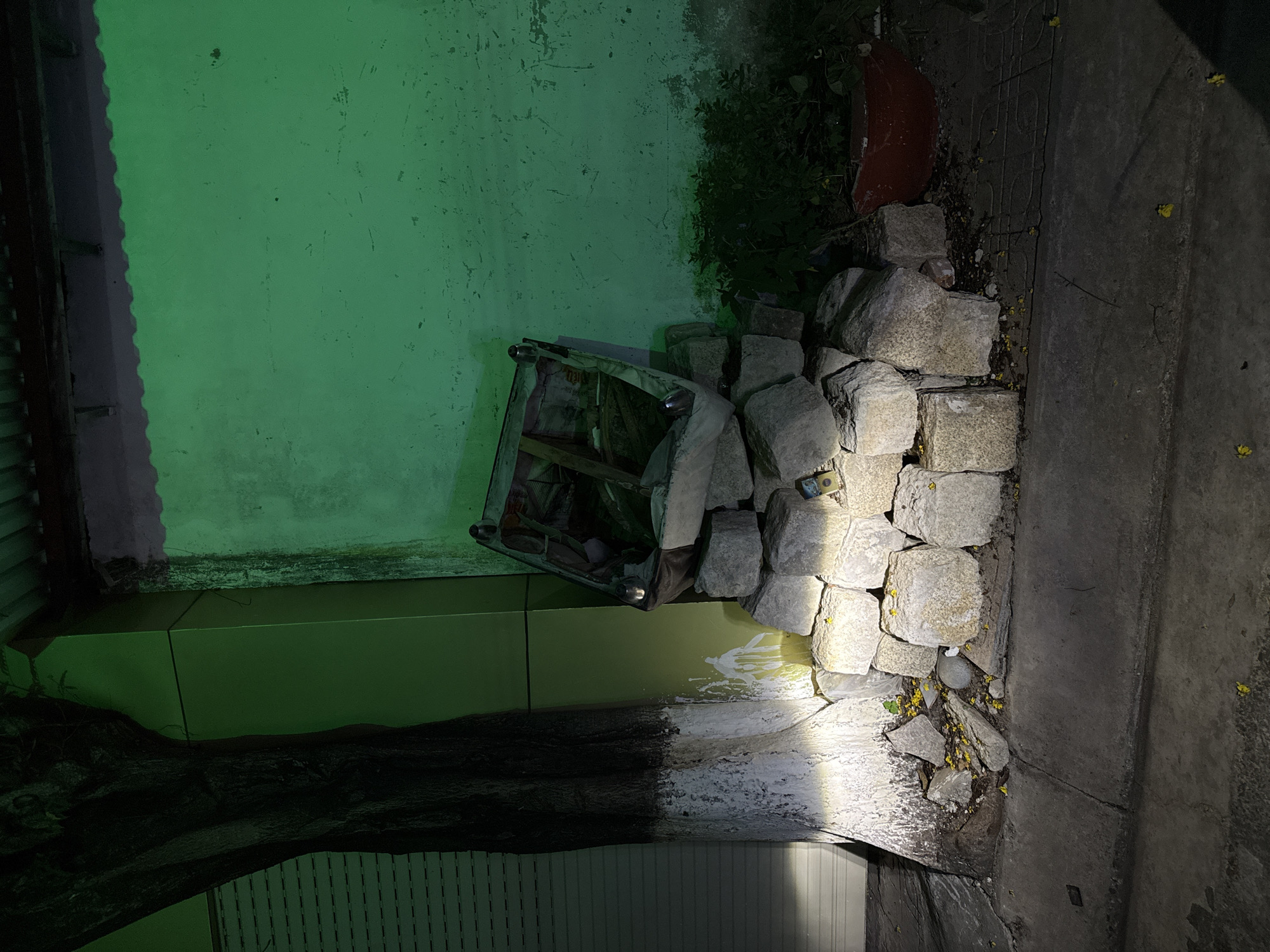An overturned, metal-framed, glass-topped table lies on its side against a pile of large, rough-hewn stones. The table appears damaged, with its glass top cracked and parts of its frame bent. Behind the table and stones, a wall painted a vibrant green is visible, with patches of wear and tear. To the left of the wall, a dark, possibly wooden structure with a corrugated edge is present. In the foreground, a rough concrete surface slopes downwards, with scattered yellow petals and debris. A tree trunk with dark bark is visible in the lower left of the image, with part of a light-colored, possibly metal-slatted structure beneath it. The scene is illuminated by a strong, artificial light source, likely a flashlight, casting harsh shadows and highlighting the textures of the stones and concrete. The time of day is indeterminate due to the artificial lighting, but the overall impression is one of disarray and neglect. There is no discernible weather or strong emotional cues. A small, yellow object, possibly a packet or sticker, is visible on top of the pile of stones. The location context is Bac Nha Trang Ward, Vietnam.