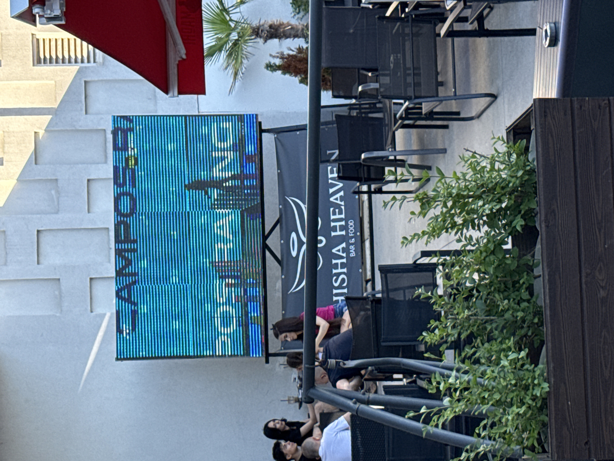 The image shows an outdoor seating area with people gathered. A large, vertical LED screen displays a pattern of blue and white vertical lines and what appears to be a silhouette of a cactus. To the right of the screen is a black banner with white text that reads "N" and "HISH HEAVEN" and "BAR & FOOD". There are several black outdoor chairs and tables arranged in the scene. Some people are seated at the tables, while others are standing or leaning. One woman in a pink top and denim shorts is visible near the banner. The scene appears to be a casual outdoor gathering, possibly at a restaurant or bar. The lighting suggests it might be daytime, with some shadows cast by the overhead structures and chairs. The overall atmosphere seems relaxed.