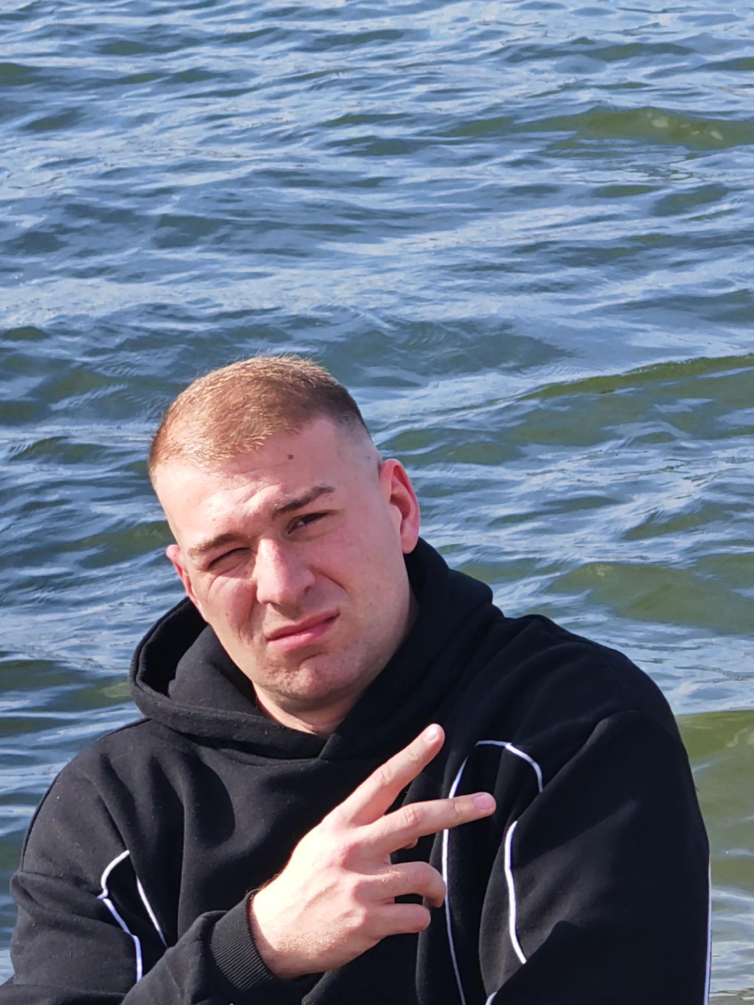 A medium shot depicts a young adult male wearing a black hooded sweatshirt with white trim. He is positioned against a backdrop of water with small waves, suggesting a body of water like a lake or sea. The man is giving a peace sign with his right hand. His hair is short and blonde. His facial expression seems slightly serious. The location, Shumen, Bulgaria, cannot be confirmed from the visual data provided.
