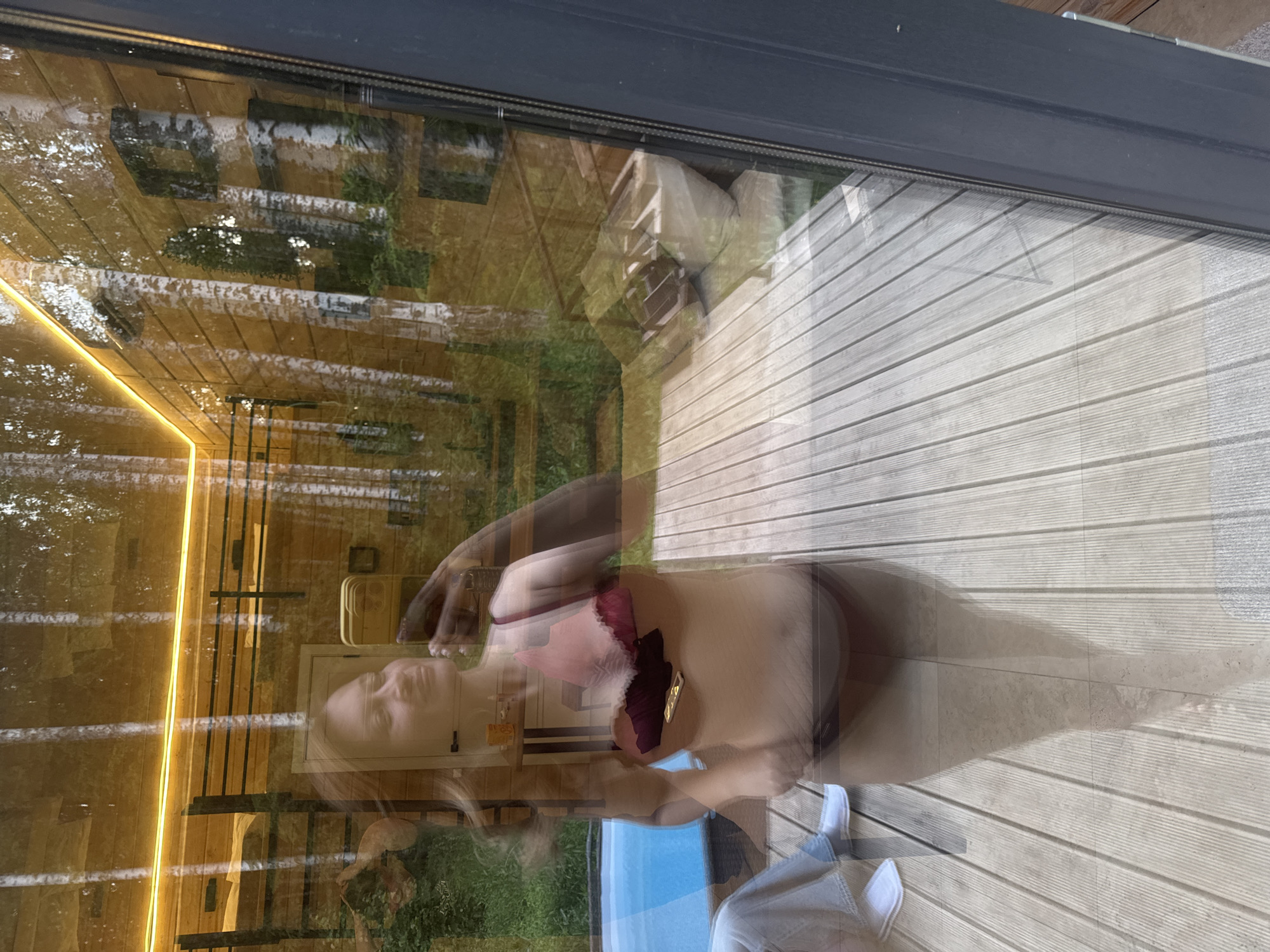 A woman is captured in a mirror selfie, wearing a bikini and holding a smartphone. The reflection shows her standing on a wooden deck with a hot tub to her left. Behind her, a wooden structure is visible, illuminated by warm lighting. The environment appears to be a natural setting, possibly a backyard or a resort area, with trees in the background. The image is taken from a high angle, looking down at the woman. There are no visible signs of the city of Bakhtiyary, Russia.