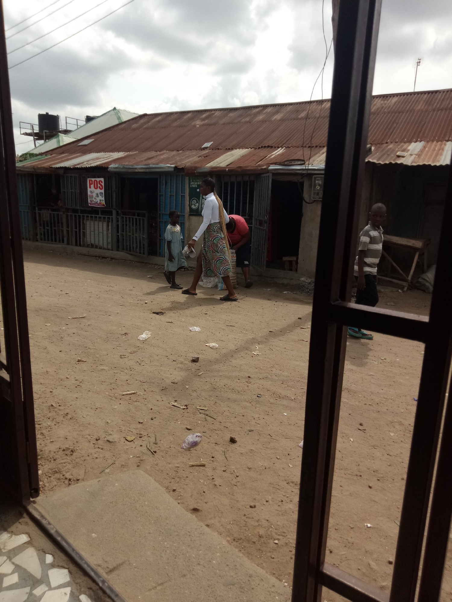 The image shows a dirt road in front of several shops with corrugated metal roofs. A young boy in a striped t-shirt and dark pants stands on the right side of the road, looking towards the camera. Further down the road, a woman in a white shirt and a patterned skirt walks away from the camera, carrying a white object. A younger child in a light blue dress walks in the same direction as the woman, slightly ahead of her. Another person in a red shirt is visible behind a metal gate to the left of the woman. The shops appear to be small businesses, with one displaying a "POS" sign. Debris, including plastic bags and leaves, is scattered on the ground. The sky is overcast with gray clouds. The scene suggests a casual street environment in a populated area.