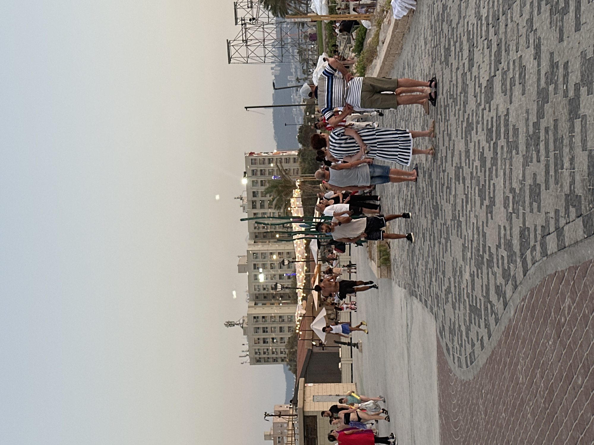 The image shows a public promenade in Kiryat Yam, Israel, likely near the beach or a waterfront area. In the background, several multi-story buildings, characteristic of urban architecture, are visible, with one prominent building featuring a cellular antenna on its roof. Two large metal structures, possibly part of an event setup or infrastructure, are also present on the right side of the frame.
The promenade is paved with a distinct pattern of grey and black rectangular stones, with a curved section of reddish-brown bricks in the foreground. Numerous people are scattered across the promenade, engaged in various activities. Several individuals are walking, some in groups, while others appear to be standing and conversing. The attire of the people suggests a warm climate, with many wearing shorts, t-shirts, and summer dresses. One person in the foreground on the right is wearing a striped t-shirt and shorts, and another individual in the center is wearing a black and white striped dress.
In the middle ground, there are some white umbrellas and what appear to be food stalls or recreational areas, indicating a lively public space. The overall atmosphere suggests a casual gathering or a leisurely stroll on a pleasant day. The sky is a pale, hazy blue, typical of a late afternoon or early evening.