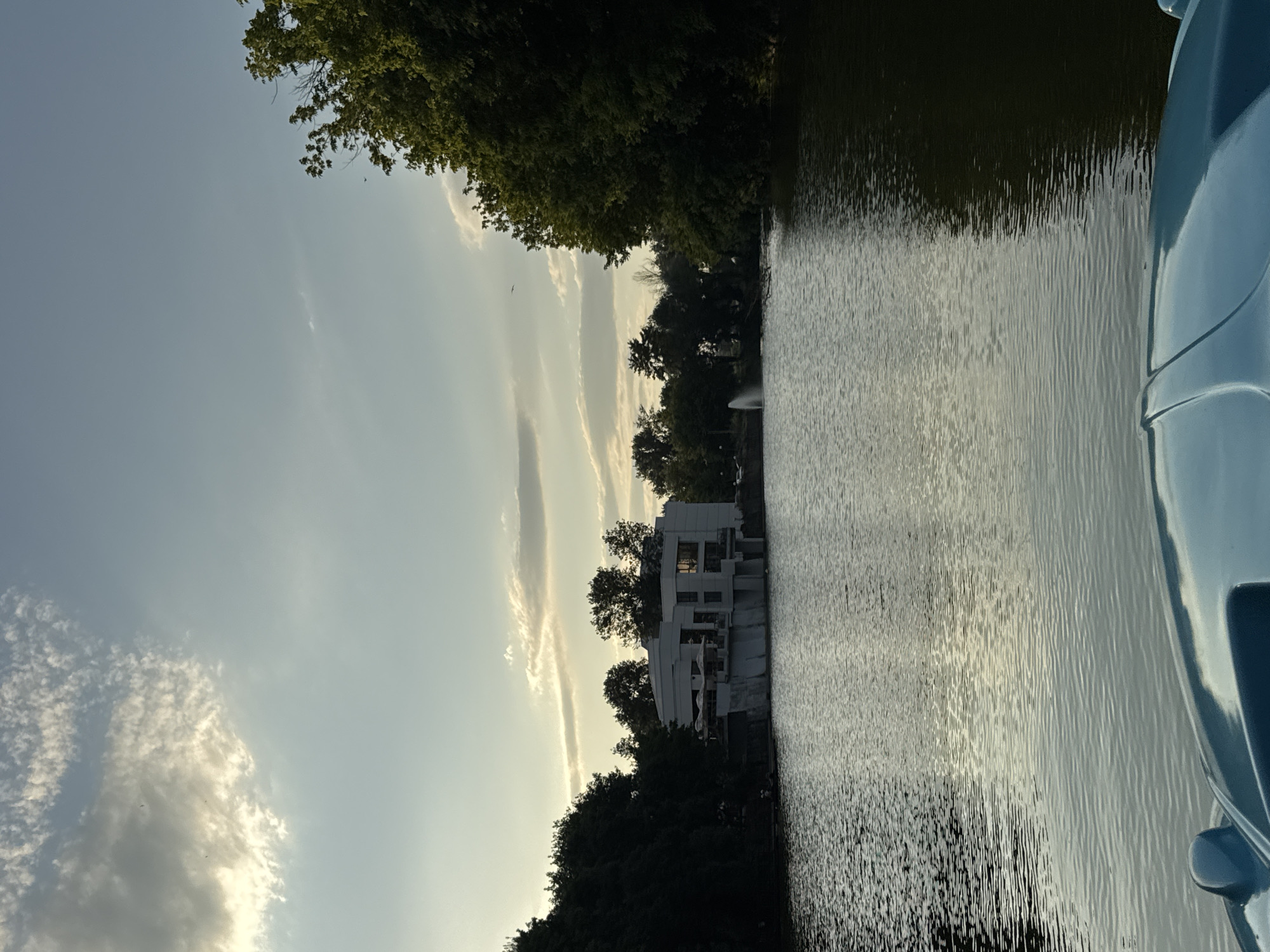 The image depicts a serene lake scene, likely within the city of Yambol, Bulgaria. In the foreground, the top of a light blue object, possibly the front of a boat or a pedal boat, is visible. The lake occupies the majority of the frame, with ripples suggesting a gentle breeze. Across the water, a modern white building with multiple levels and large windows stands on the shore, surrounded by lush green trees. A fountain sprays water into the air in the distance. The sky above is a pale blue, with scattered white clouds, illuminated by the soft light of what appears to be late afternoon or early morning. The trees on either side of the lake are dense and green, adding to the natural beauty of the setting. The overall impression is one of tranquility and calm.