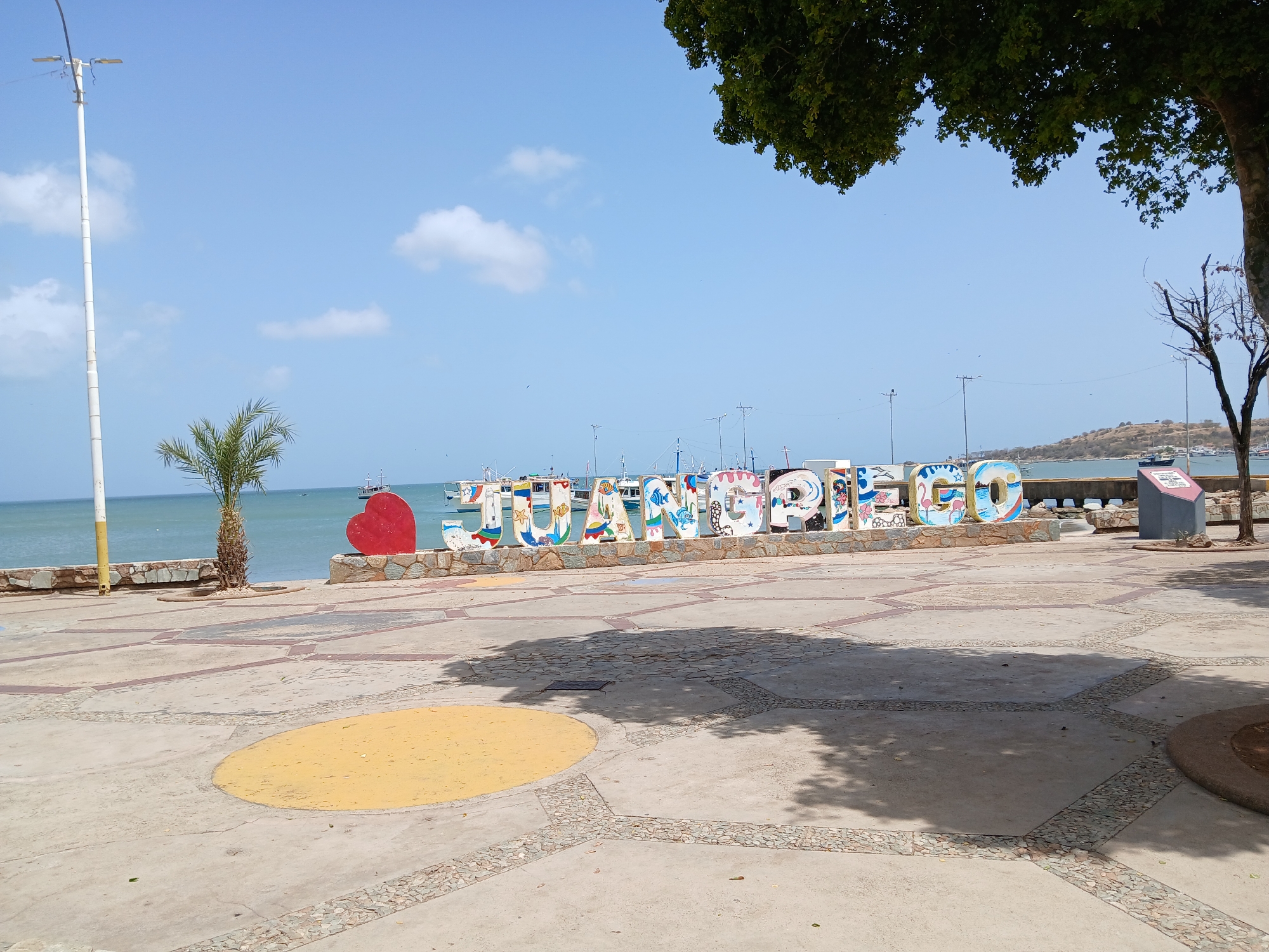 The image depicts a sunny day at a waterfront location in Juan Griego, Venezuela, showcasing a large, colorful "JUAN GRIEGO" sign. The sign is comprised of individual letter-shaped structures, each uniquely painted with vibrant designs. The sign is situated on a low stone wall or platform, overlooking the sea. A palm tree stands to the left of the sign. Various boats are visible on the water in the distance. The foreground features a paved public space with a pattern of large concrete tiles and stone accents. A large tree casts a shadow on the pavement. In the right side of the frame is a sign with unreadable text.