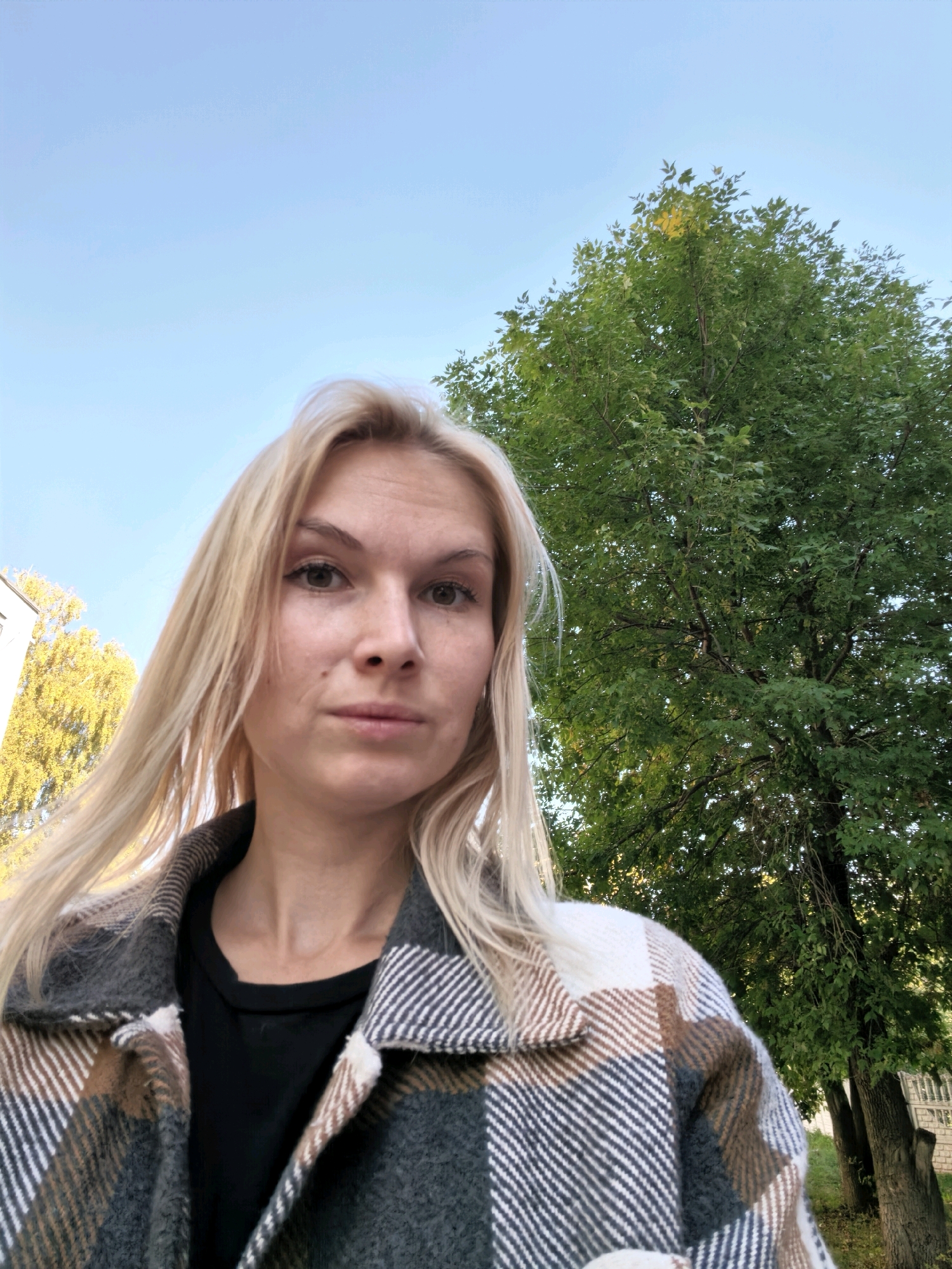 A medium-shot, slightly low-angle selfie captures a young blonde woman in Sterlitamak, Russia. She is wearing a black t-shirt beneath a plaid jacket with brown, white, and gray squares. Her blonde hair is partially obscuring her face, which is turned slightly to her left. Her expression is neutral, with her eyes looking directly at the camera.
The background features a bright blue sky and the dense green foliage of a large tree to the right, with its trunk visible. To the left of the woman, a building with yellowing leaves on a tree in front of it is partially visible. The overall lighting suggests it is daytime.