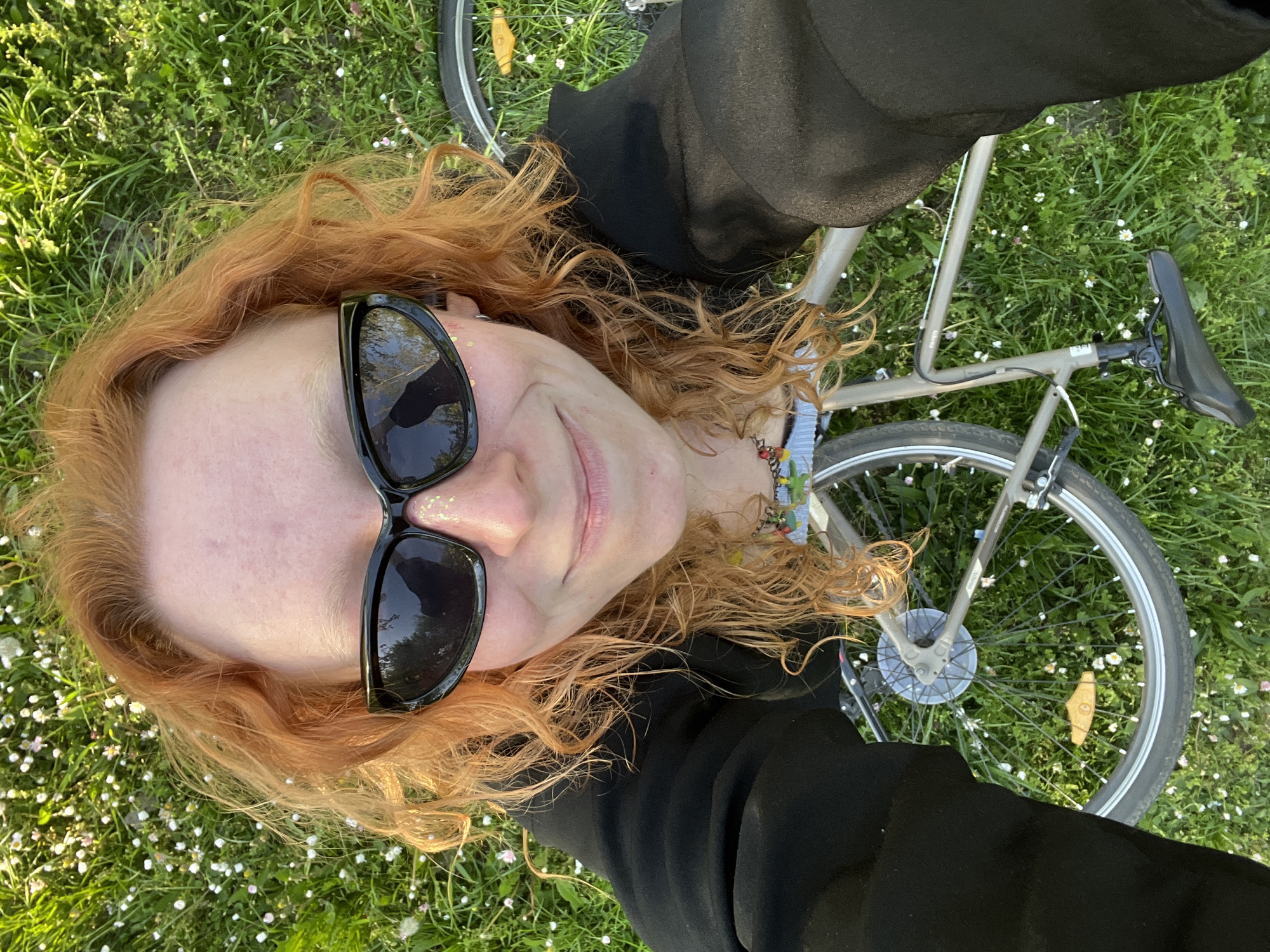 An overhead shot captures a person with long, wavy reddish-orange hair lying on their back in a grassy area. They wear black sunglasses that cover their eyes, and a slight smile is visible on their lips. Small golden glitter or flakes are applied near their temple and cheekbone. The person is dressed in a dark, likely black, long-sleeved top. A bicycle, with a silver-grey frame and black saddle, is positioned behind and to the side of the person, partially obscured by their body and arm. A decorative, colorful beaded bracelet is visible on their wrist. The setting is a lush green lawn or field, dotted with numerous small white and light pink flowers, suggesting a spring or summer day. The scene is brightly lit by natural sunlight, indicating either morning or late afternoon. The overall impression is one of relaxation outdoors. No legible text is visible in the image. The photo was taken in Cukarica Urban Municipality, Serbia.