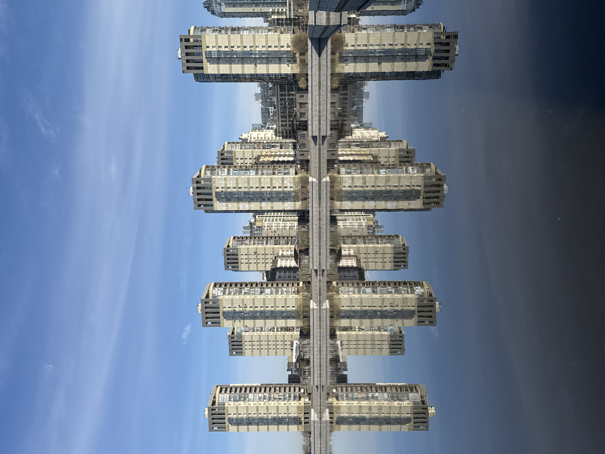 The image shows a reflection of buildings in water, with the sky visible above and below. The buildings appear to be modern residential structures, possibly apartment complexes, constructed from light-colored materials. A walkway or road appears to be dividing the real building and its reflection. Based on the architectural style and location of the photo, it could be in the city of Saint Petersburg, Russia.