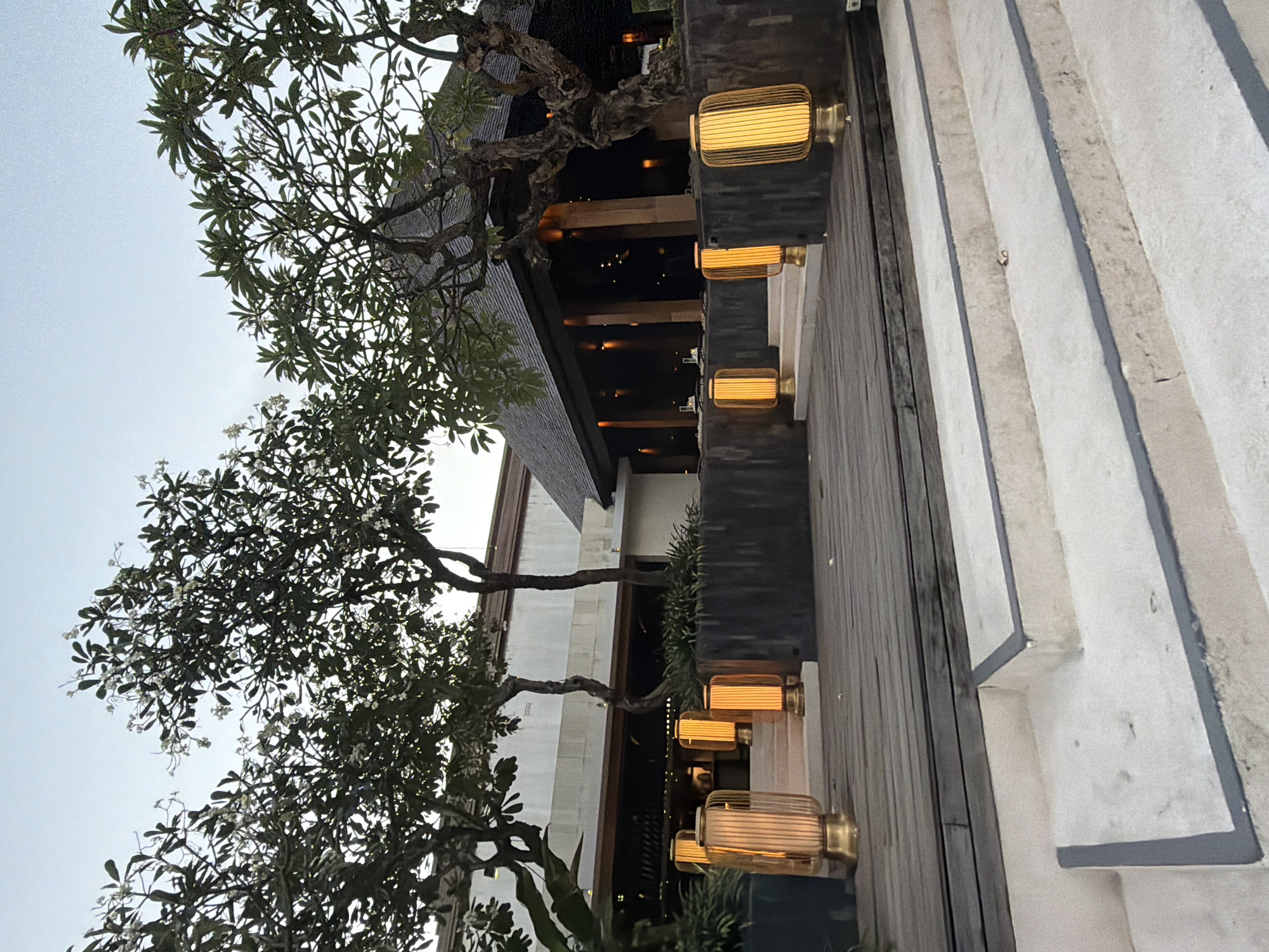 The image shows an exterior view of a building with traditional Balinese architecture. The building features a dark, thatched roof and an open-air dining area with warm lighting. Several tall, cylindrical lanterns with a golden hue illuminate a wooden walkway and steps leading to the building's entrance. Lush green trees with delicate white flowers are visible on either side of the structure, framing the scene. The sky is overcast, suggesting it might be dusk or a cloudy day. The location is a resort or restaurant in Kuta, Indonesia, likely a high-end establishment given the architecture and landscaping. There are no people or animals visible in the image, and no actions or interactions are taking place. The scene is clean and well-maintained, with a focus on creating an inviting and tranquil atmosphere.