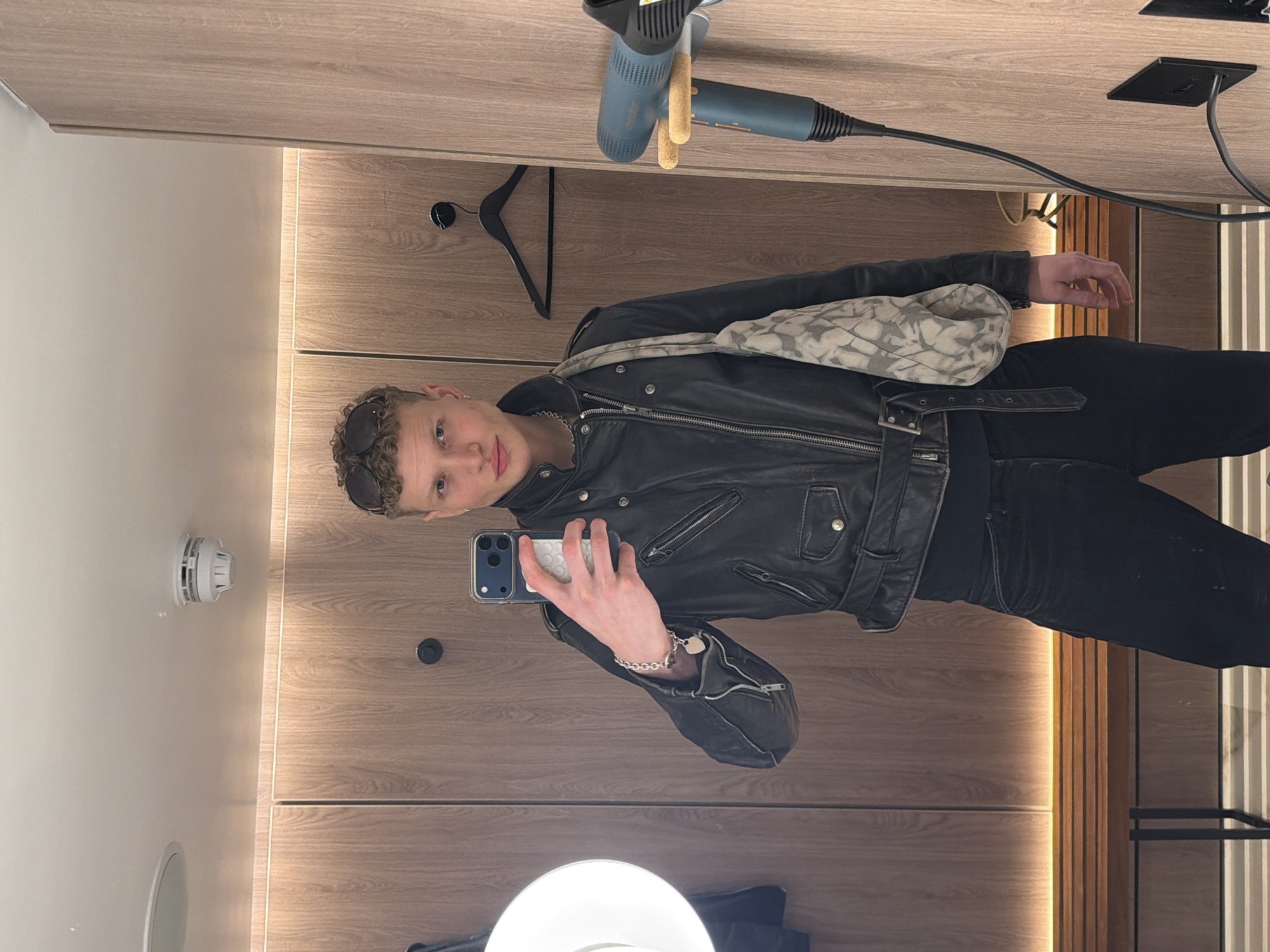 A young individual with short, curly light brown hair and blue eyes, wearing black sunglasses on their head, a black leather motorcycle jacket, black undershirt, and black jeans, is taking a mirror selfie. Their left wrist, adorned with a silver bracelet featuring a heart charm, holds a blue smartphone with a white textured case. A silver chain necklace is visible around their neck, and a light grey, patterned shoulder bag is draped over their right shoulder. The scene is a modern, minimalist interior, indicative of a hotel room or changing area in London, United Kingdom. The background consists of light wood-paneled walls, an integrated slatted wooden bench, and a light-colored ceiling. Overhead, a circular ceiling light fixture and a white smoke detector are present. On the right wall, a black coat hanger and a mounted blue Valera Swiss Made hairdryer, plugged into a black wall outlet, are visible.