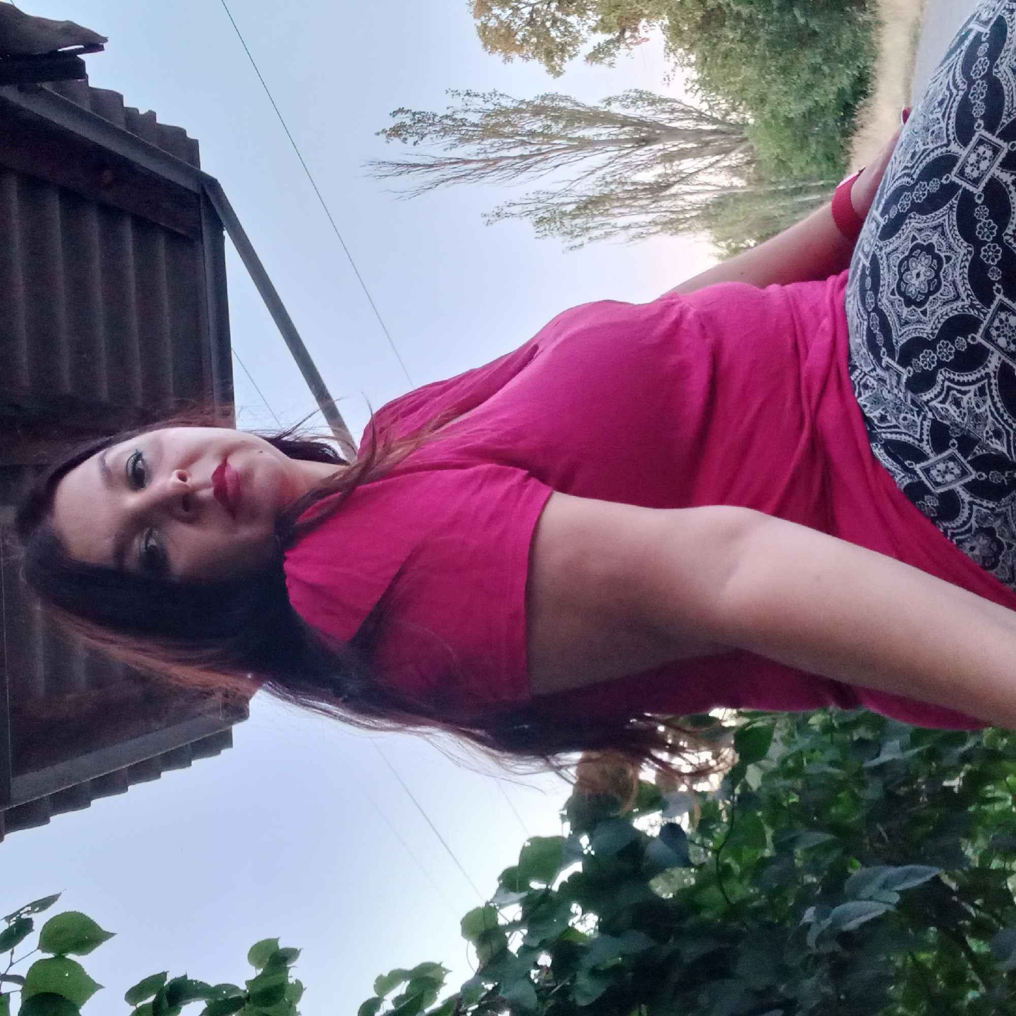 The image captures a woman seated outdoors, with a clear sky and lush greenery in the background. She is wearing a vibrant pink t-shirt and patterned pants. Her hair is dark and long, and she is looking towards the camera with a neutral expression. The woman is positioned in front of a covered structure, possibly a gazebo or a simple roof, supported by metal beams. A cluster of green leaves is visible on the left side of the frame, framing the woman. In the distance, several tall, slender trees stand against the sky, adding to the natural backdrop. The overall scene suggests a relaxed outdoor setting. The specific location in Berezhynka, Ukraine, is not discernible from the visual elements present in the image.