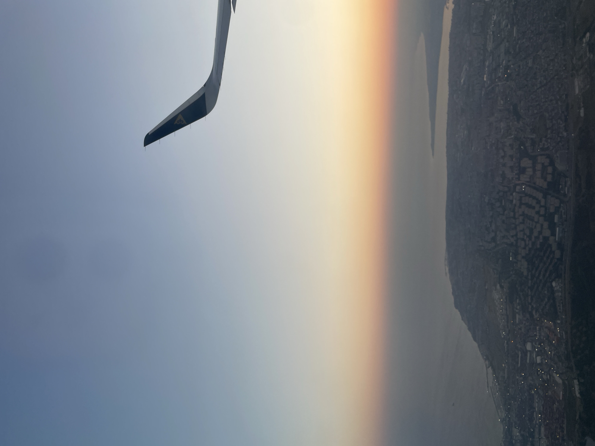 An aerial view captures the city of Beykoz, Turkey, under a gradient sky of pale blue transitioning to a soft orange hue near the horizon, indicative of either sunrise or sunset. The foreground is dominated by the wing of an airplane, its tip visible in the upper right quadrant, featuring a dark stripe with a yellow insignia. Below the wing, the sprawling urban landscape of Beykoz unfolds, densely populated with buildings. The city is situated along the coastline of a body of water, likely the Bosphorus Strait. Numerous lights are visible within the city, suggesting it is either dusk or dawn. The overall scene conveys a sense of travel and a panoramic perspective of a coastal city.