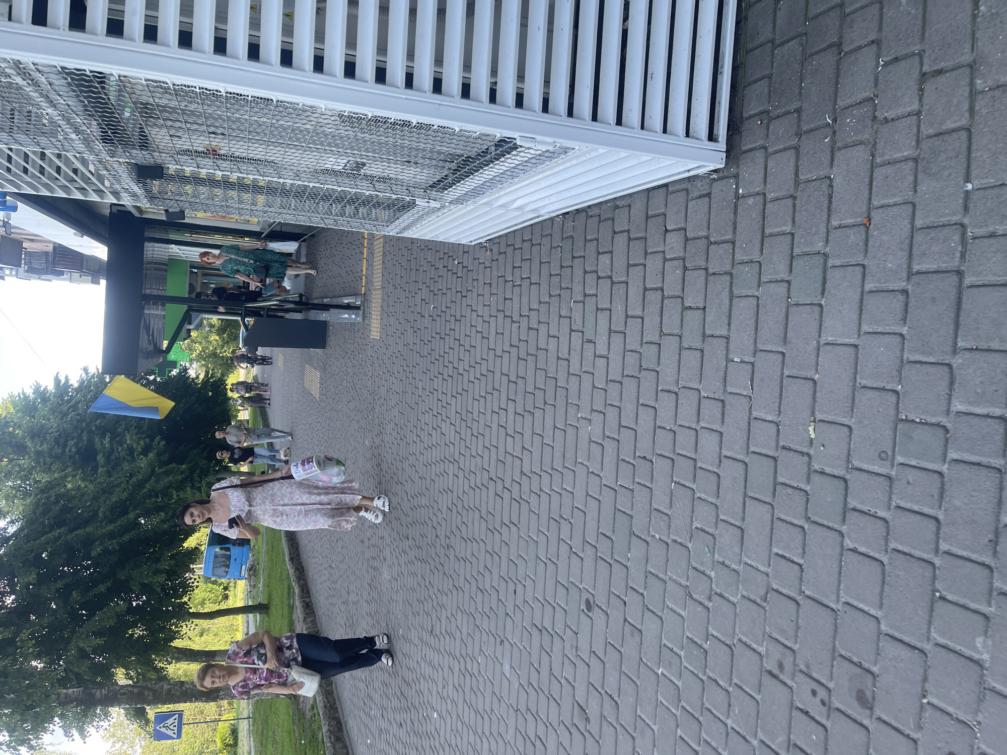 A medium shot captures a pedestrian walkway in an urban setting, with a focus on a few individuals walking. The walkway is paved with grey interlocking bricks. On the left side of the image, a woman with short brown hair, wearing a floral-patterned short-sleeved top and dark pants, walks with a white shoulder bag. To her right, a woman in a white floral dress with short sleeves and white sandals is walking and holding a shopping bag in her left hand, while using her smartphone with her right hand. Behind them, a blue van is parked on the side of the road, and a Ukrainian flag is visible on a pole in the background. The right side of the image shows the exterior of a building with a white louvered structure and a metal grate. A woman in a green dress is seen walking away from the building entrance. The scene appears to be outdoors during the daytime with sunlight casting shadows.