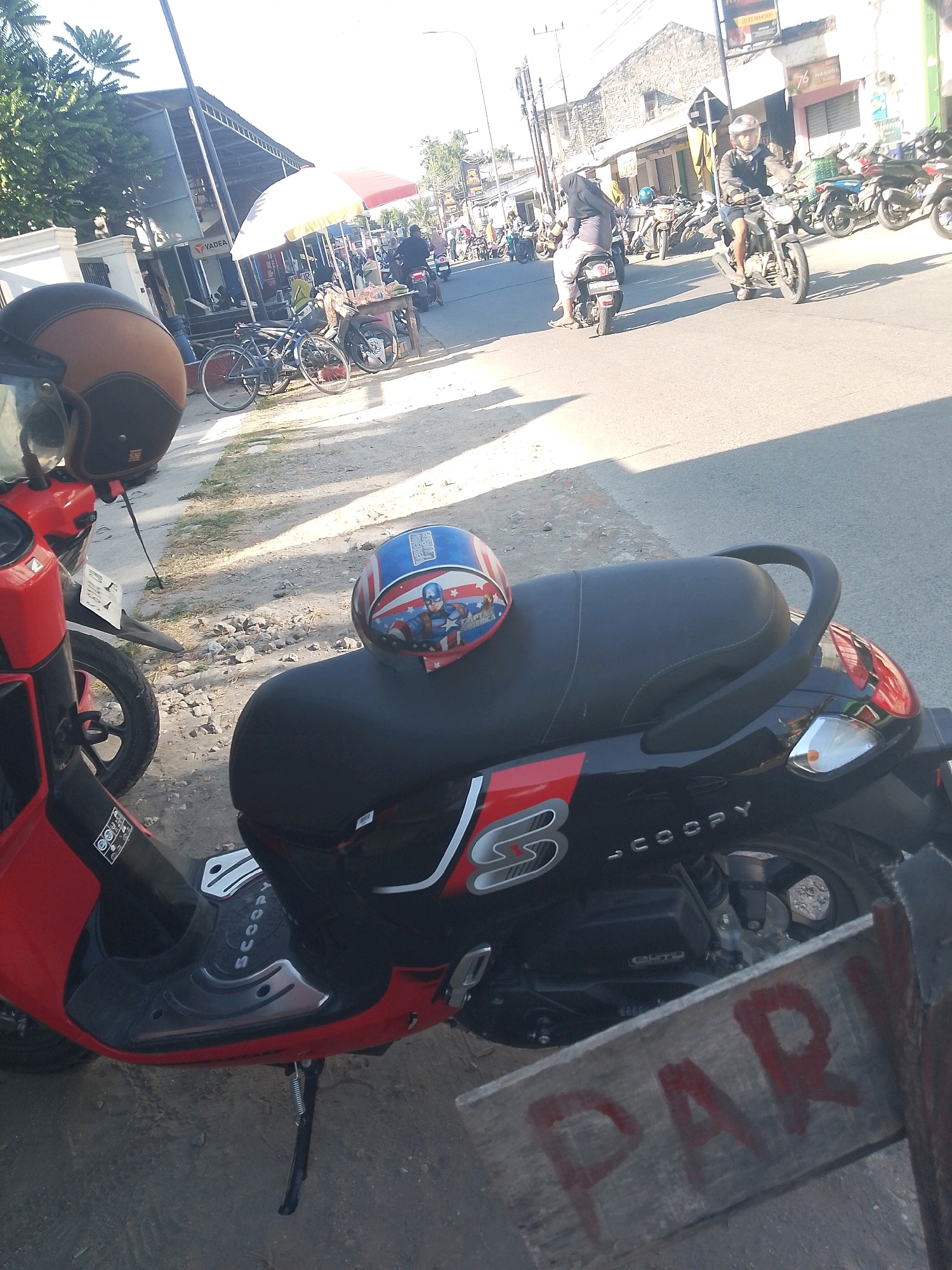 A street scene in Jampet, Indonesia, features several motorcycles and scooters parked and in motion. A red and black scooter is prominently in the foreground, displaying the "SCOOPY" logo. A helmet with a Captain America design rests on the scooter's seat. In the background, a variety of other motorcycles are visible, and a few individuals are seen riding them. Buildings line the street, and a yellow and red umbrella shades a vendor stall. The overall impression is of a busy, sunny day with everyday street activity.
