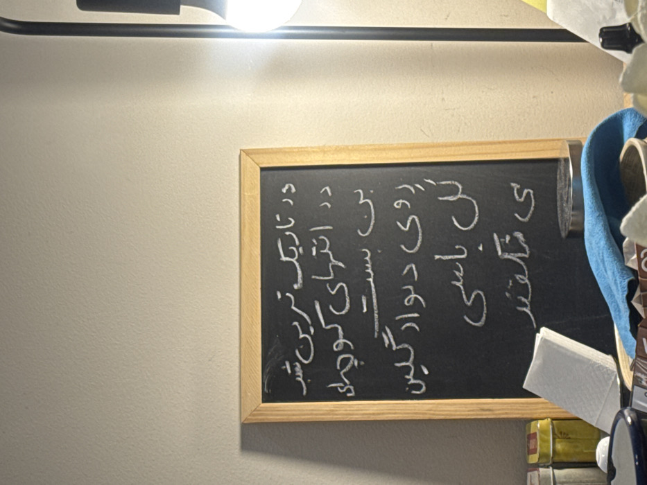 This image depicts an indoor scene located in Milan, Italy. A light-colored wall serves as the background for a rectangular blackboard with a light wooden frame, centrally positioned. The blackboard features text written in white chalk, rendered in Persian script. To the immediate right, a black lamp stand with an illuminated, exposed light bulb provides direct illumination to the area. A flat surface beneath the blackboard holds various objects. From left to right, these include a partially visible yellow container with a red lid, a stack of white paper napkins, and a blue item (possibly a bowl or ashtray). Further right, a blue fabric cloth partially obscures a silver circular object and a brown container. To the far right, a pile of irregular, light-colored crystalline material and a small, dark bottle with a dropper cap are visible. No individuals are present in the scene.