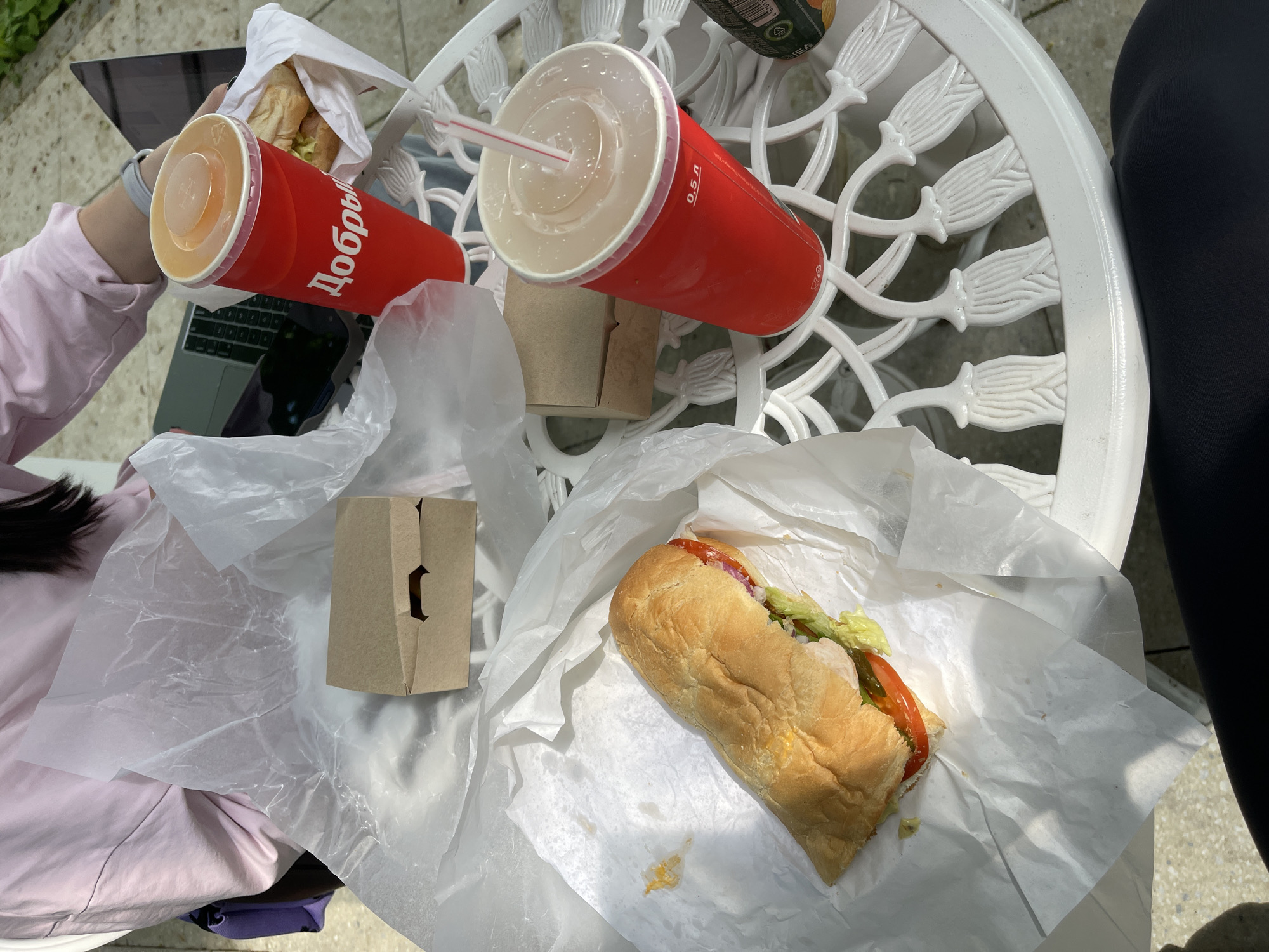 On a sunny day in Moscow, Russia, a light-skinned person with dark hair and wearing a light pink long-sleeved shirt sits at a white wrought-iron table outdoors, with a laptop nearby. Two sandwiches wrapped in white paper, one in hand, and two large red paper cups with lids and straws sit on the table along with two small brown paper bags. One cup is labeled "Добрый" (Dobry). A black legging-covered leg is partially visible near the edge of the table. A can of dark-colored soda is partially visible on the table.