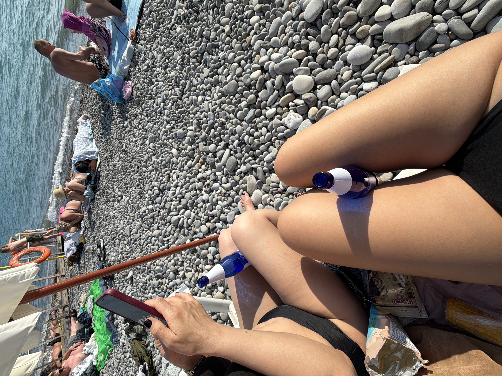 The image depicts a sunny day at a pebble beach in Mamedova Shchel', Russia. In the foreground, two individuals are seated on the pebbled shore, with their legs extended. One individual, on the left, is wearing a black swimsuit and holding a mobile phone. A blue bottle sits on the lap of the person on the right, also dressed in black swimwear. In the background, several other beachgoers are visible, some lying on towels and others standing near the water's edge. The sea is calm and blue, with gentle waves lapping the shore. Beach umbrellas provide shade for some of the sunbathers. Several bags and personal items are scattered around the beach. A lifebuoy is visible attached to a structure in the background. The overall scene conveys a relaxed atmosphere of a typical summer day at the seaside.