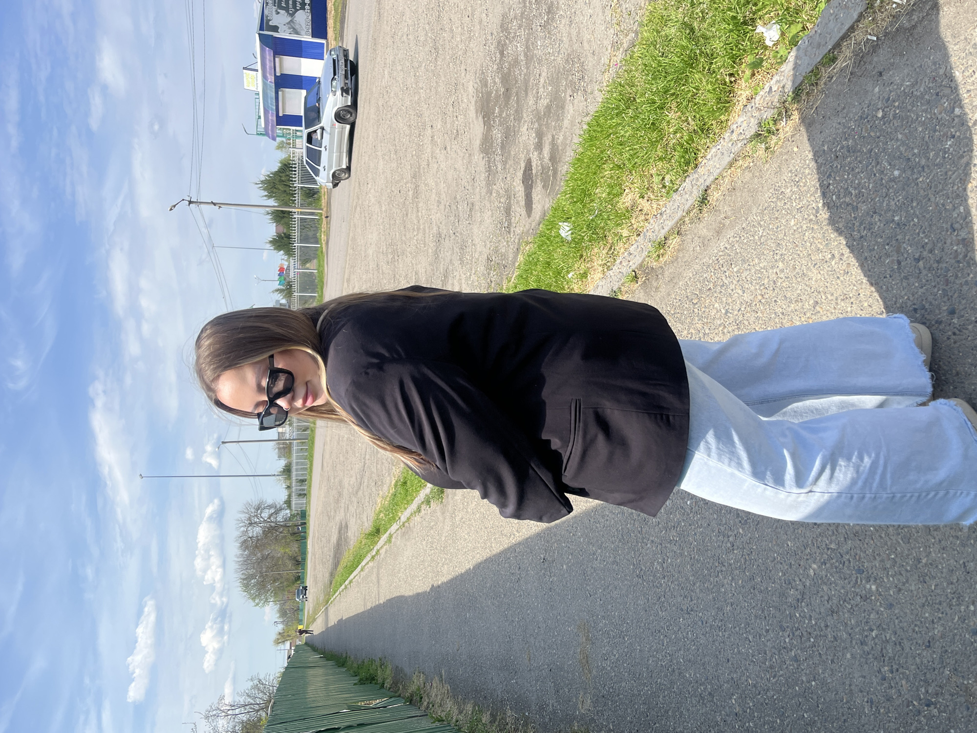 A young woman in sunglasses and a black blazer stands on a grey asphalt path in Novopokrovskaya, Russia. She wears light blue jeans. A green fence lines one side of the path, while grass and a concrete curb border the other. In the background, utility poles and scattered trees are visible against a partly cloudy sky. A building and vehicles are farther in the distance.