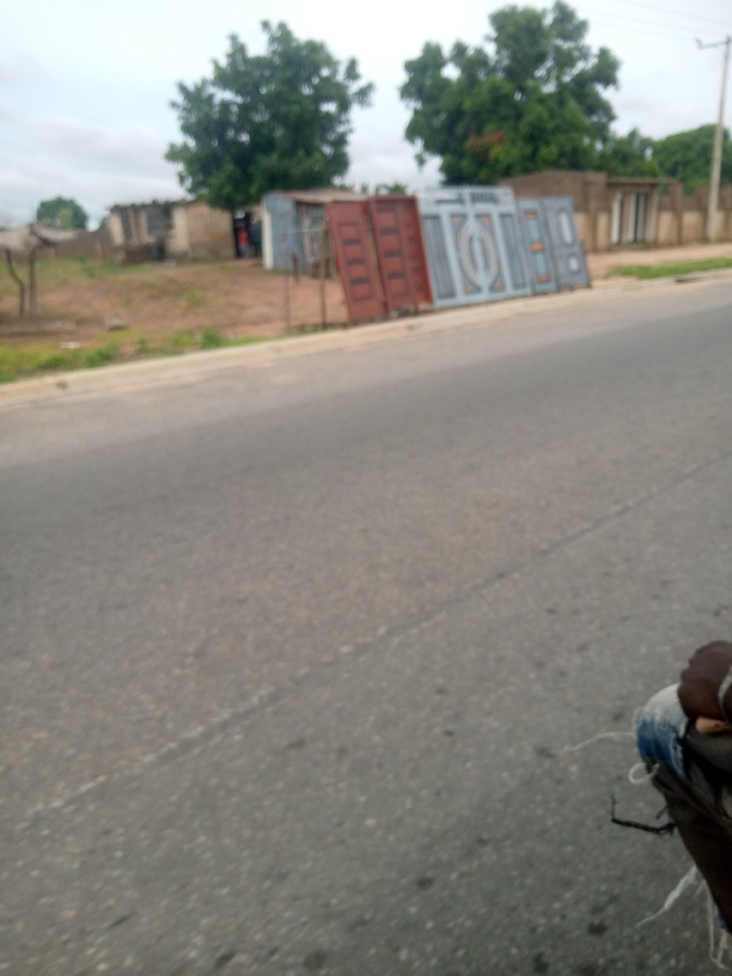 The image depicts a street scene in Jalingo, Nigeria, featuring several metal gates propped up along the roadside. These gates appear to be for sale or display, with a variety of designs including intricate patterns and solid panels. In the background, a building with a concrete wall and a simple structure with a corrugated metal roof are visible, suggesting a commercial or residential area. A few trees are scattered throughout the scene, with dense foliage indicating a possibly humid climate. The road itself is asphalt and appears to be a two-lane street. The overall impression is of a busy, informal marketplace or roadside vendor area.