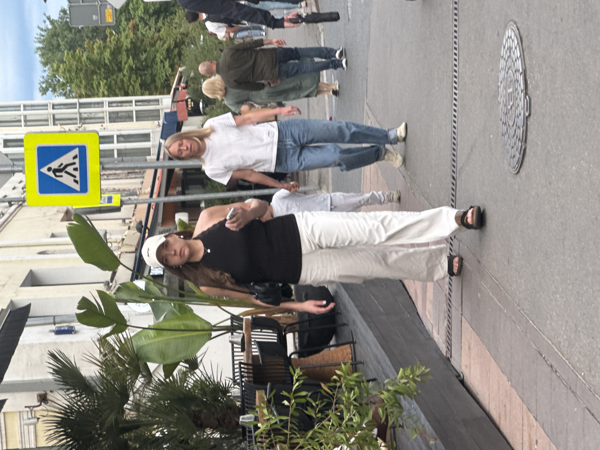 The image shows a street scene with several people. The scene is oriented vertically, making it appear as if the people are walking on the ceiling. In the foreground, a young woman with long brown hair, wearing a white baseball cap, black tank top, and white pants, is holding a phone and looking at it. She is standing next to a large potted plant with broad green leaves. Behind her, another person in a white t-shirt and blue jeans is visible, also looking down. Further in the background, several other individuals are walking along the sidewalk. One person is wearing a dark green jacket and blue jeans, and another has blonde hair. A yellow pedestrian crossing sign is prominently displayed on the left side of the image. The background shows buildings with windows and trees, suggesting an urban environment. The lighting suggests it is daytime. A manhole cover with a circular pattern is visible on the asphalt. The overall impression is of people casually walking on a street on a pleasant day.