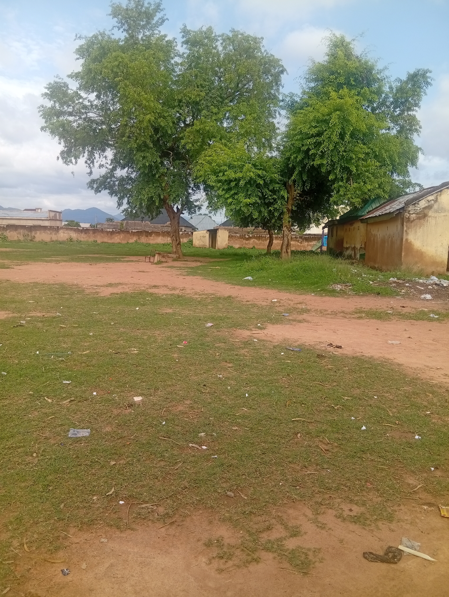 The image depicts an outdoor, rural or semi-urban scene under a partly cloudy sky in Unknown, Unknown. The foreground and midground consist of an unpaved, reddish-brown dirt field with numerous patches of green grass and scattered small litter, including plastic bags and wrappers. A prominent dirt path traverses the ground from the lower right towards the center. In the midground, several large, mature trees with dense green foliage stand, one to the left and a cluster to the right. Under the left tree, a small, rudimentary wooden bench or structure is visible. In the background on the right, two weathered, single-story buildings with light brown/yellow walls and corrugated metal roofs are present. Behind the field on the left, a long, faded orange-brown wall separates the area from other residential structures with diverse roof styles, and a distant mountain range is visible against the horizon. No distinct human figures or animals are clearly identifiable. The lighting indicates daylight conditions.