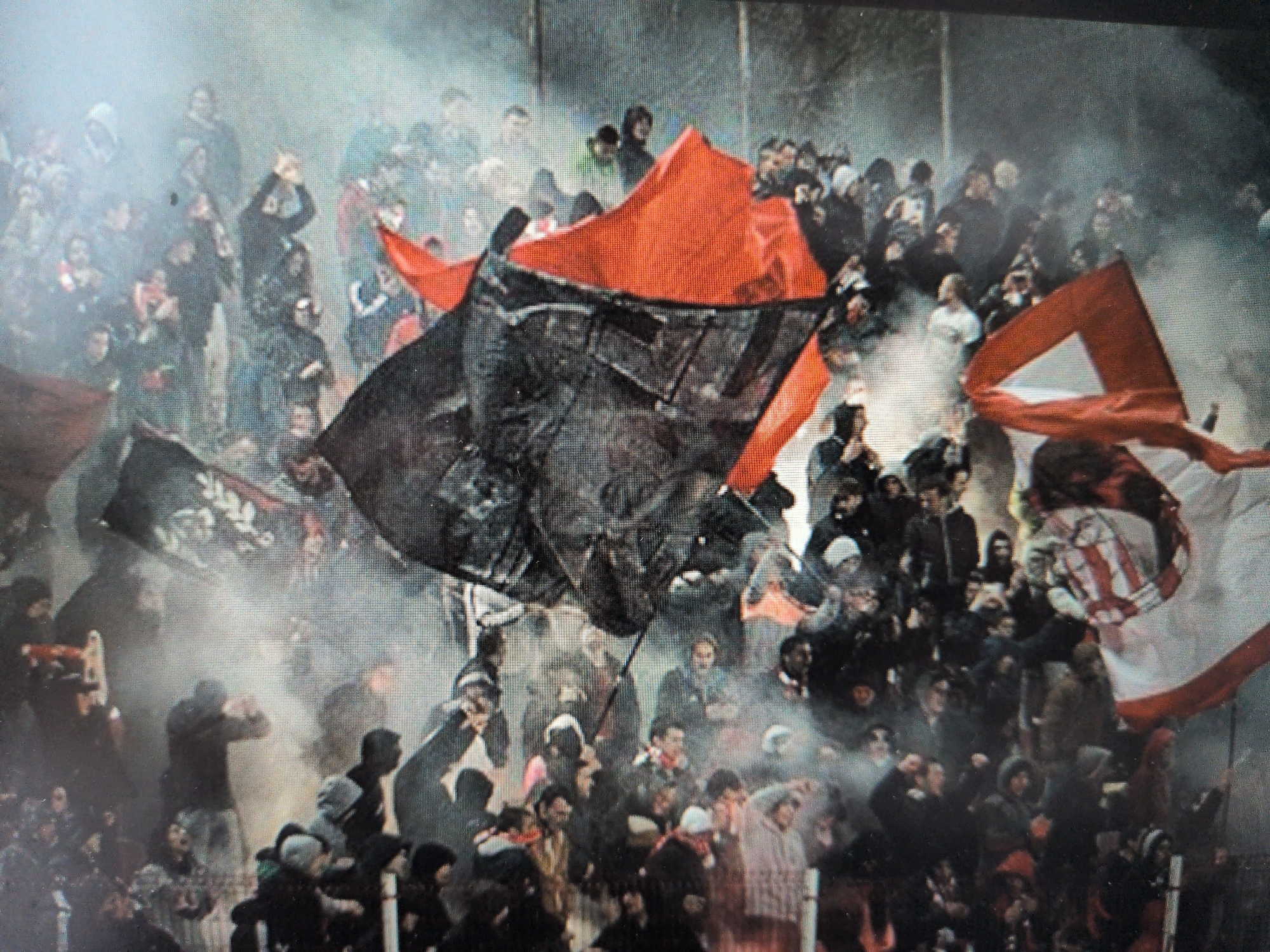 The image captures a dense crowd of people, likely sports fans, in an energetic and hazy atmosphere. The scene is dominated by a multitude of individuals packed closely together, many with their arms raised or gesturing. The lighting is somewhat dim and obscured by a significant amount of smoke or fog, which permeates the entire frame.
Several large flags are prominent objects in the foreground and midground. There are multiple bright red flags, some billowing upwards, adding strong splashes of color. A large black flag, appearing worn or textured, with some indistinct white markings or designs, is central in the midground. Another notable flag, visible on the right side, is striped in red and white, possibly featuring a crest or emblem, though details are unclear due to motion and blur.
The activity suggests a passionate gathering, likely at a sporting event such as a football match, given the prevalence of supporter culture indicated by the flags and smoke. The crowd appears animated, possibly cheering, chanting, or reacting to events. The smoke could originate from flares or smoke bombs, common at such events, contributing to the intense and somewhat chaotic ambiance.
Notable details include the widespread smoke obscuring much of the background and softening the outlines of individuals. The faces of the people are mostly indistinct due to distance, resolution, and the smoky conditions, but their collective energy is palpable. There is no clearly legible text on any of the flags or visible surfaces. The setting appears to be an outdoor stadium or arena, possibly a terraced section. The time of day is hard to ascertain due to the artificial lighting and atmospheric conditions, but it could be late afternoon or evening.