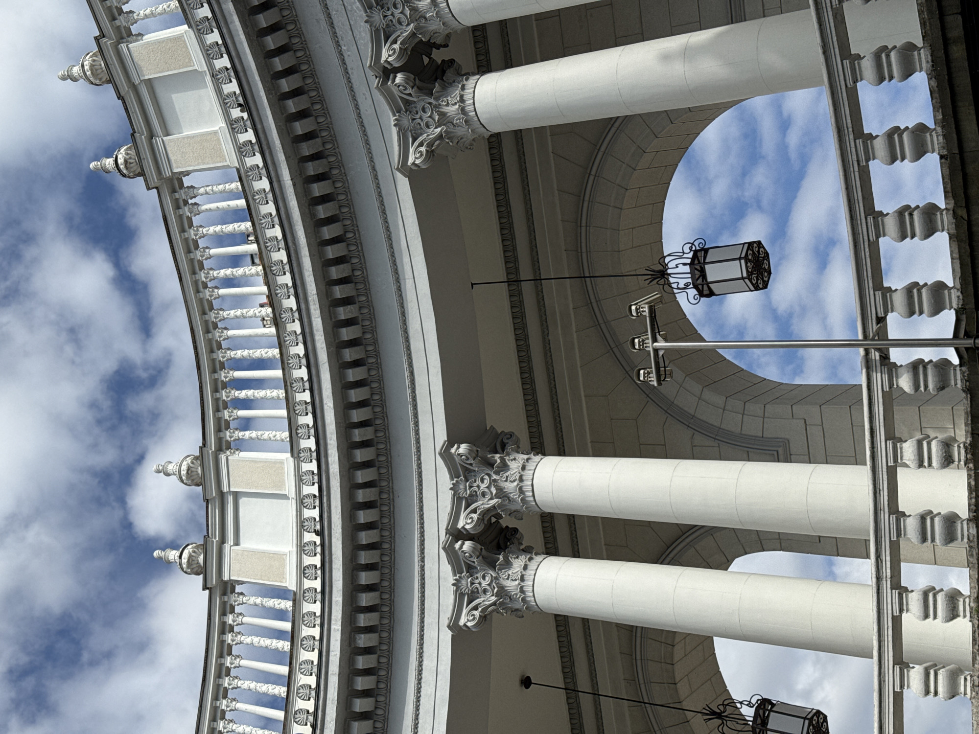 The image shows an upward view of an architectural structure in Sochi, Russia. The structure features classical elements such as white columns with ornate capitals, a curved balcony with decorative railings, and an arched opening revealing a blue sky with clouds. Hanging from the structure are black, lantern-style light fixtures. The color scheme is predominantly white and gray, characteristic of classical architecture.
