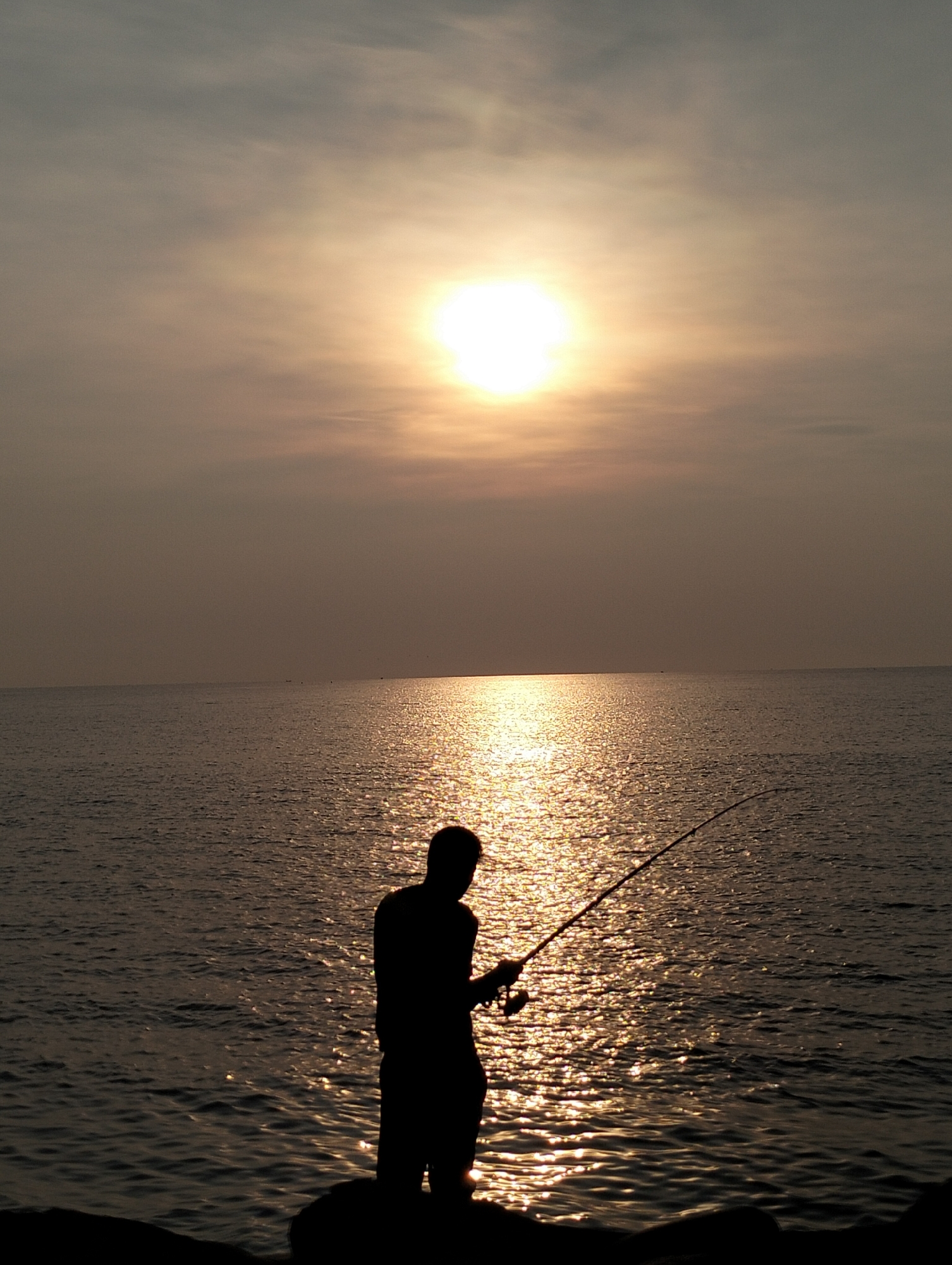 A silhouette of a person is seen fishing on a shore, with the sun setting over the sea. The sun is reflecting off the water, creating a shimmering effect. The location is possibly in Lhokseumawe, Indonesia, given the prompt. The silhouette suggests it is either early morning or late afternoon due to the angle of the sun. The person is holding a fishing rod, casting it out into the water. The environment appears to be a coastal area. There are no clear interactions evident in the picture, only the action of fishing.