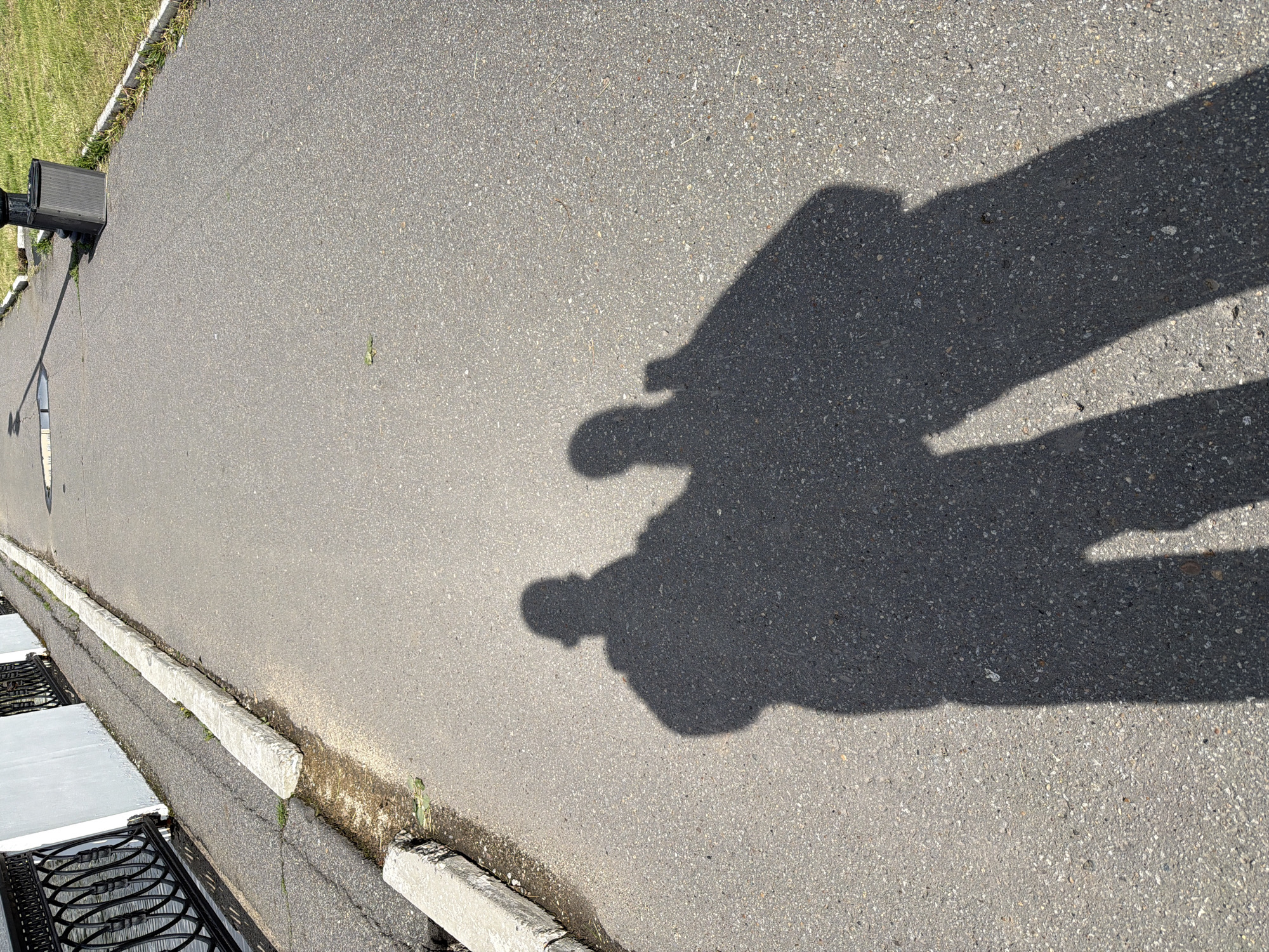 The image depicts an asphalt surface, likely a road or parking area, with two distinct human shadows cast upon it. The shadows suggest two individuals standing close together. A sidewalk runs along the left edge of the frame, delineated by a concrete curb. Adjacent to the sidewalk is a metal grate covering a drainage area. In the background, a grassy area is visible. The image provides no specific information to identify the location as Izhevsk, Russia.