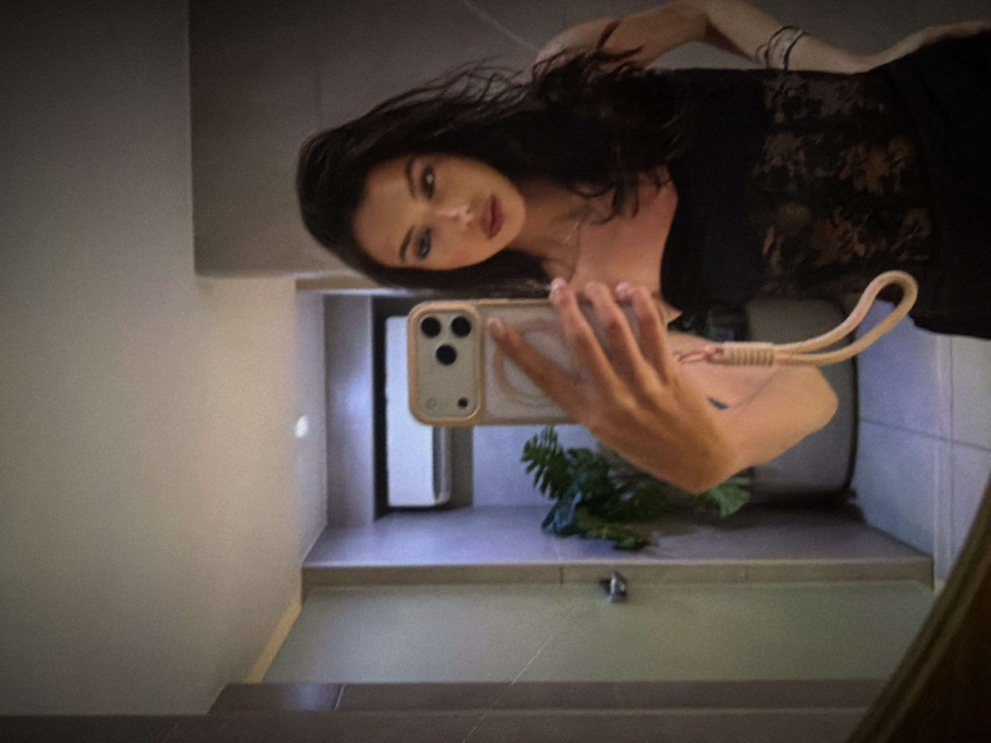 A woman with long, dark, wavy hair takes a selfie in a bathroom. She is wearing a black strapless top with lace detailing and a necklace. Her right hand, holding a phone with a light-colored case and strap, is visible in the foreground. In the reflection of the mirror, a door, a potted plant, and an air conditioning unit are visible. The floor is tiled.