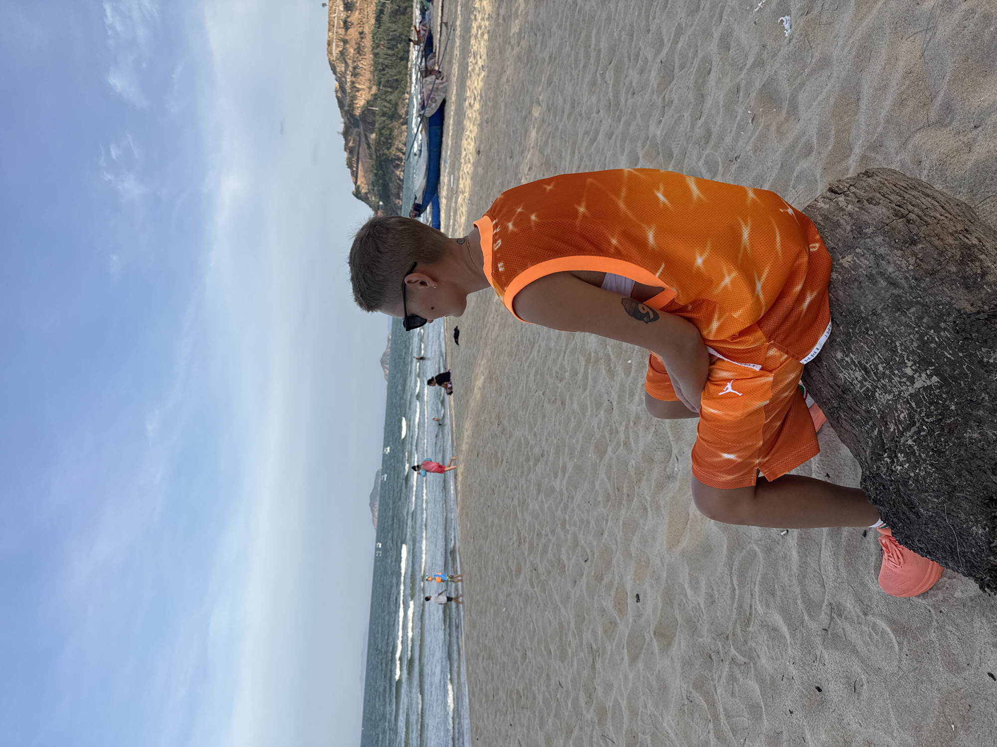 A person with short hair and sunglasses is sitting on a piece of driftwood on a sandy beach, facing the ocean. They are wearing an orange tie-dye tank top over a white sports bra and orange shorts with white trim. The person has a tattoo on their left arm. In the background, the ocean has waves, and people are visible walking and playing on the beach. Boats are docked on the sand further down the coast, and a hilly, vegetated landscape is visible in the distance. The sky is mostly clear with some scattered clouds. This scene is located on the beach in South Quy Nhon Ward, Vietnam.