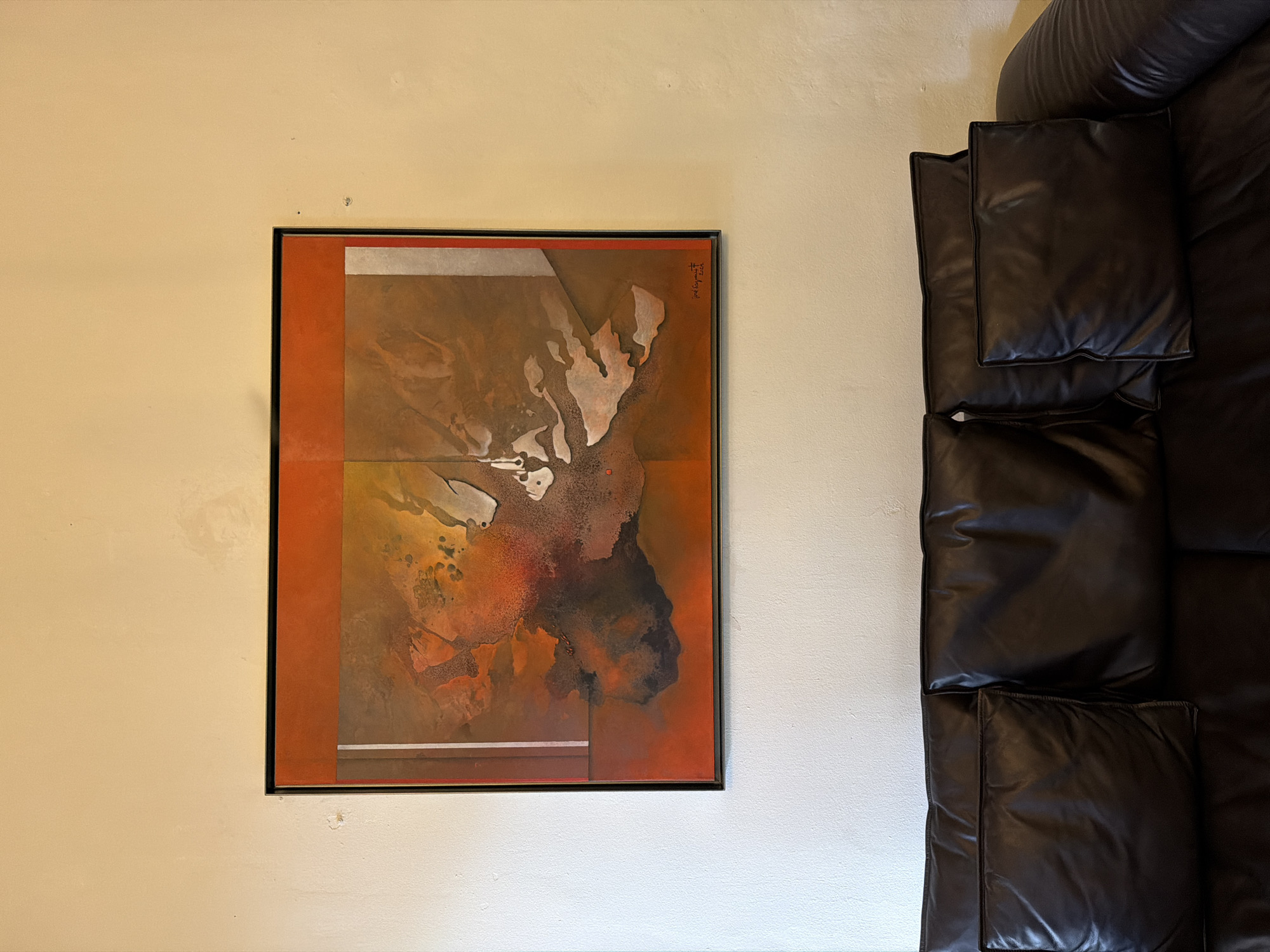 A framed painting hangs on a beige wall next to a dark brown leather sofa. The painting is predominantly orange and brown, with a central abstract design featuring lighter, possibly white or silver, elements. The painting is framed in black. The leather sofa has large, square cushions. The wall is plain and textured. The location is not specified in the image and cannot be determined.
