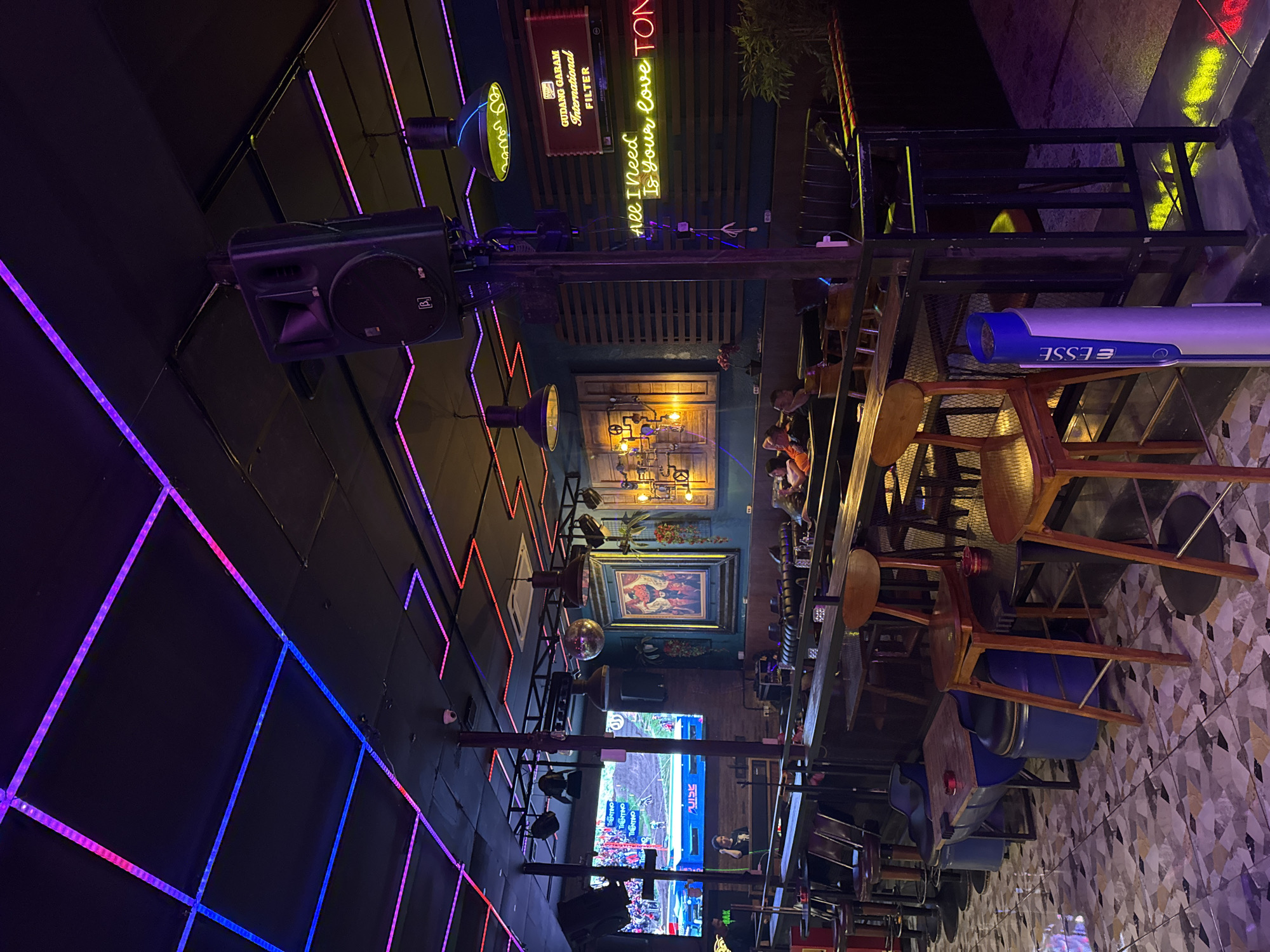 This image depicts the interior of a bar in Kuta, Indonesia, characterized by its vibrant neon lighting and modern, industrial aesthetic. The ceiling is adorned with a network of blue and pink LED strip lights, creating a dynamic and atmospheric glow. A large speaker is mounted on a stand, suggesting a space designed for music and entertainment.
The bar area features high-top tables and stools, with some patrons seated in the background. The decor includes artistic elements such as framed artwork and decorative lighting fixtures, contributing to a trendy and inviting ambiance. The floor is tiled with a mosaic pattern, and a prominent neon sign reads "All I Need Is Your Love TON."
In the background, a large screen displays an image of a motocross event, indicating that the bar may also serve as a venue for watching sports. The overall impression is that of a lively establishment that blends contemporary design with entertainment features.