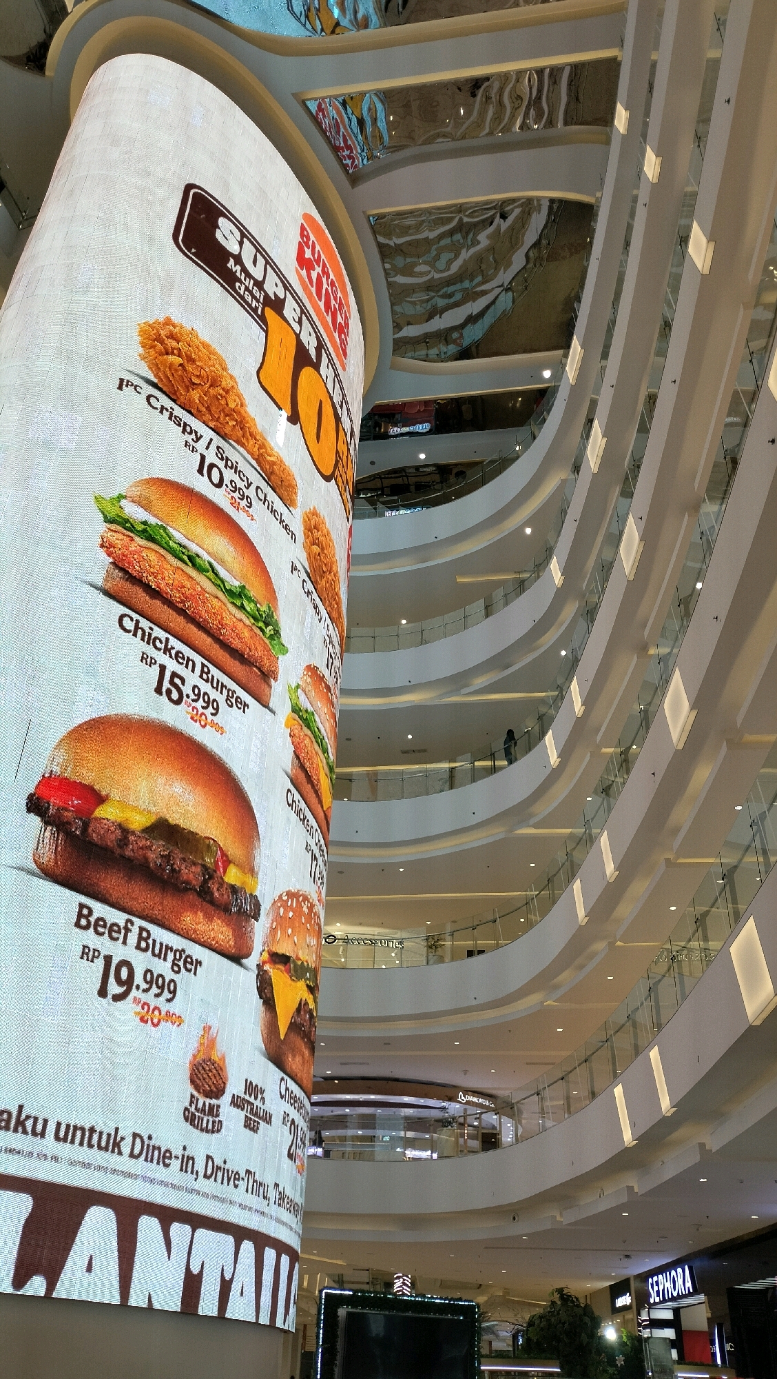The image shows a large, curved LED screen displaying a Burger King advertisement inside a multi-level shopping mall. The advertisement features pictures of chicken burgers, beef burgers, and fried chicken, along with prices in Indonesian Rupiah (RP). The mall features modern architecture with white curved balconies and glass railings, illuminated by interior lighting. Reflections of the mall's interior and ceiling can be seen on the curved surfaces of the upper levels. In the background, several shops are visible on different floors, including Sephora. The overall scene is well-lit and suggests a commercial environment.